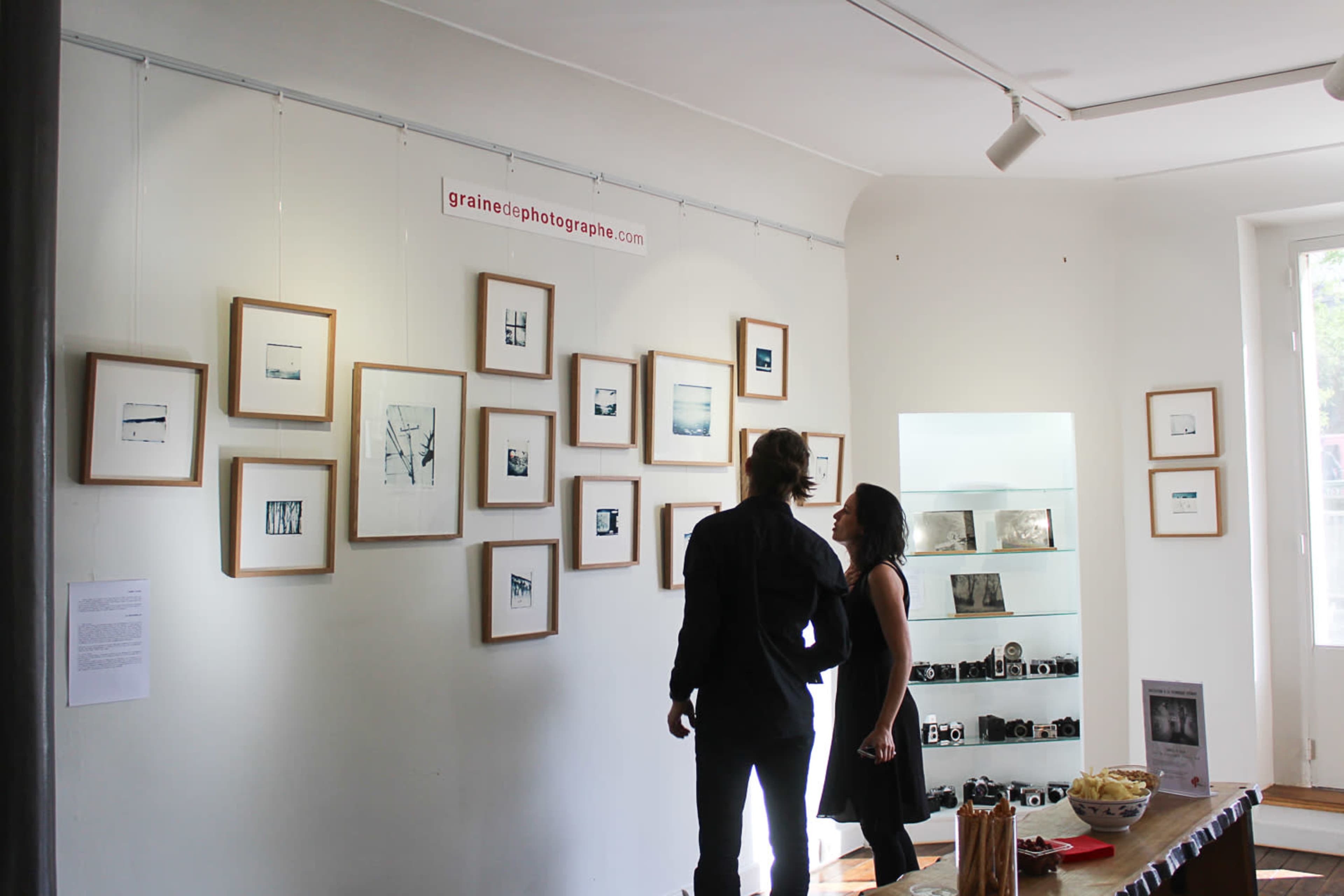 Two people examine framed photographs displayed on a gallery wall, while a shelf with cameras and light snacks is visible nearby.