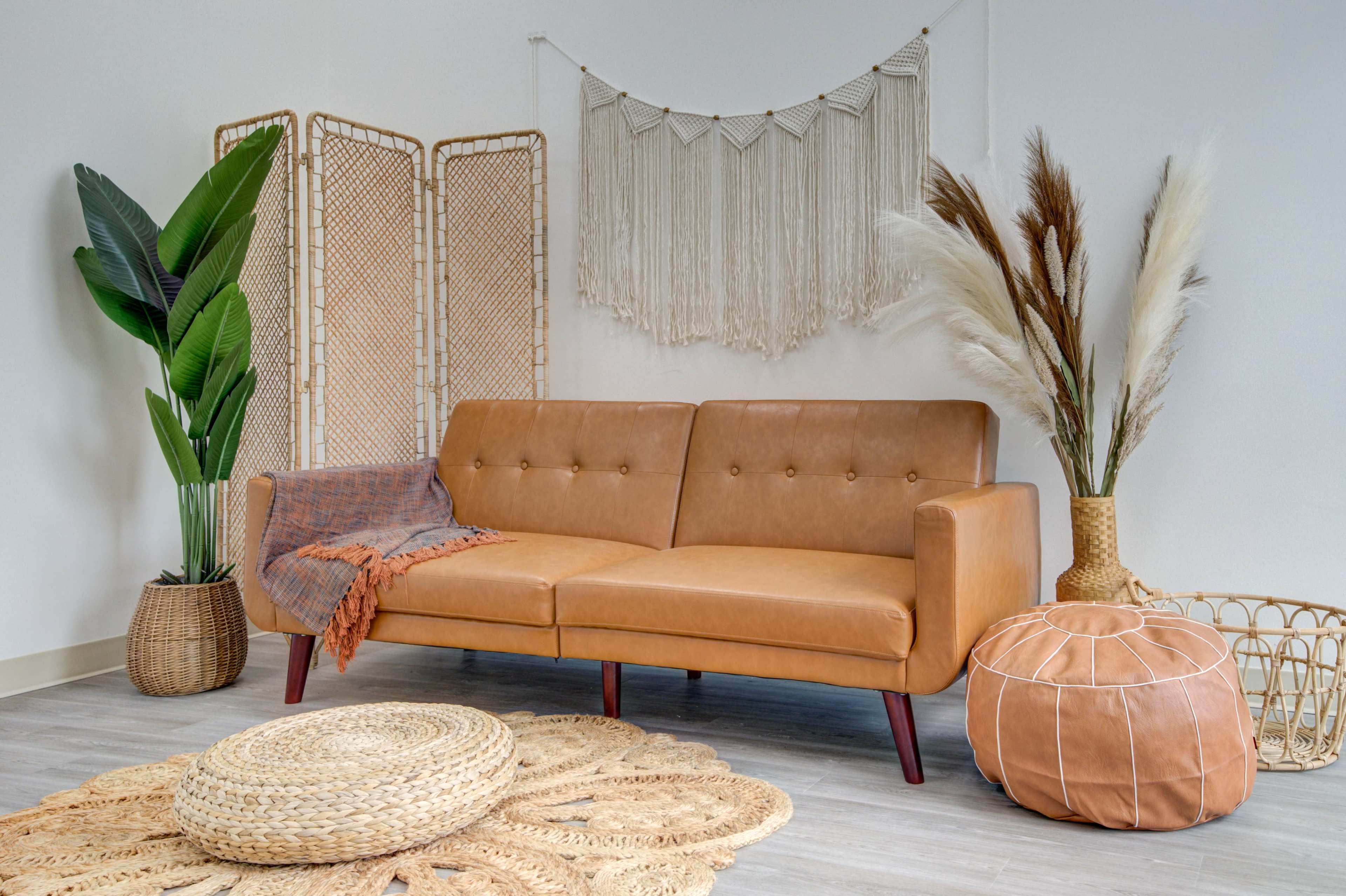 A brown leather sofa is positioned in a light-colored room, surrounded by decorative plants, a macramé wall hanging, and various woven decor items.
