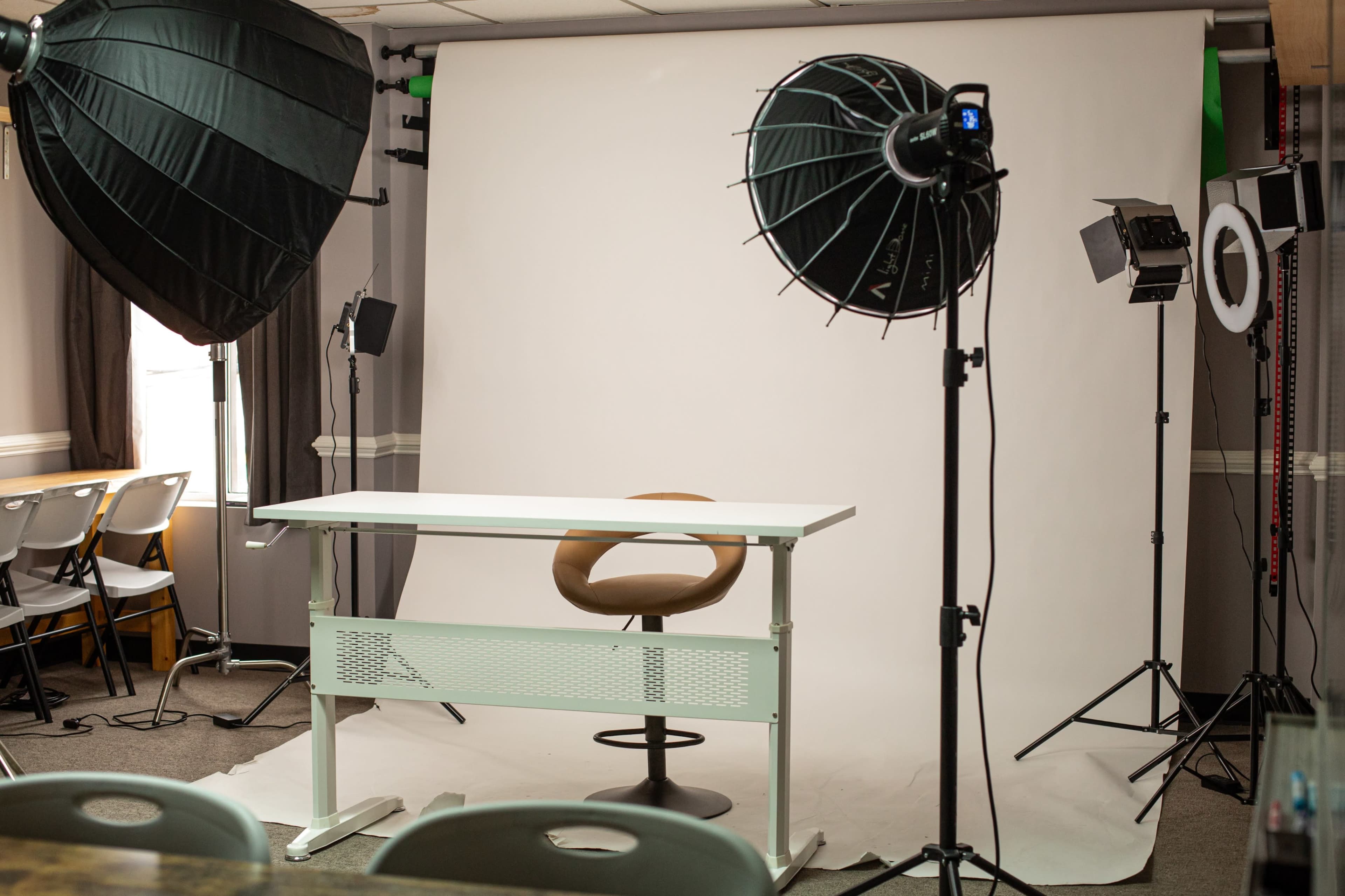 The image shows a photography studio setup with a plain backdrop, a chair, a table, and various lighting equipment positioned around the scene.