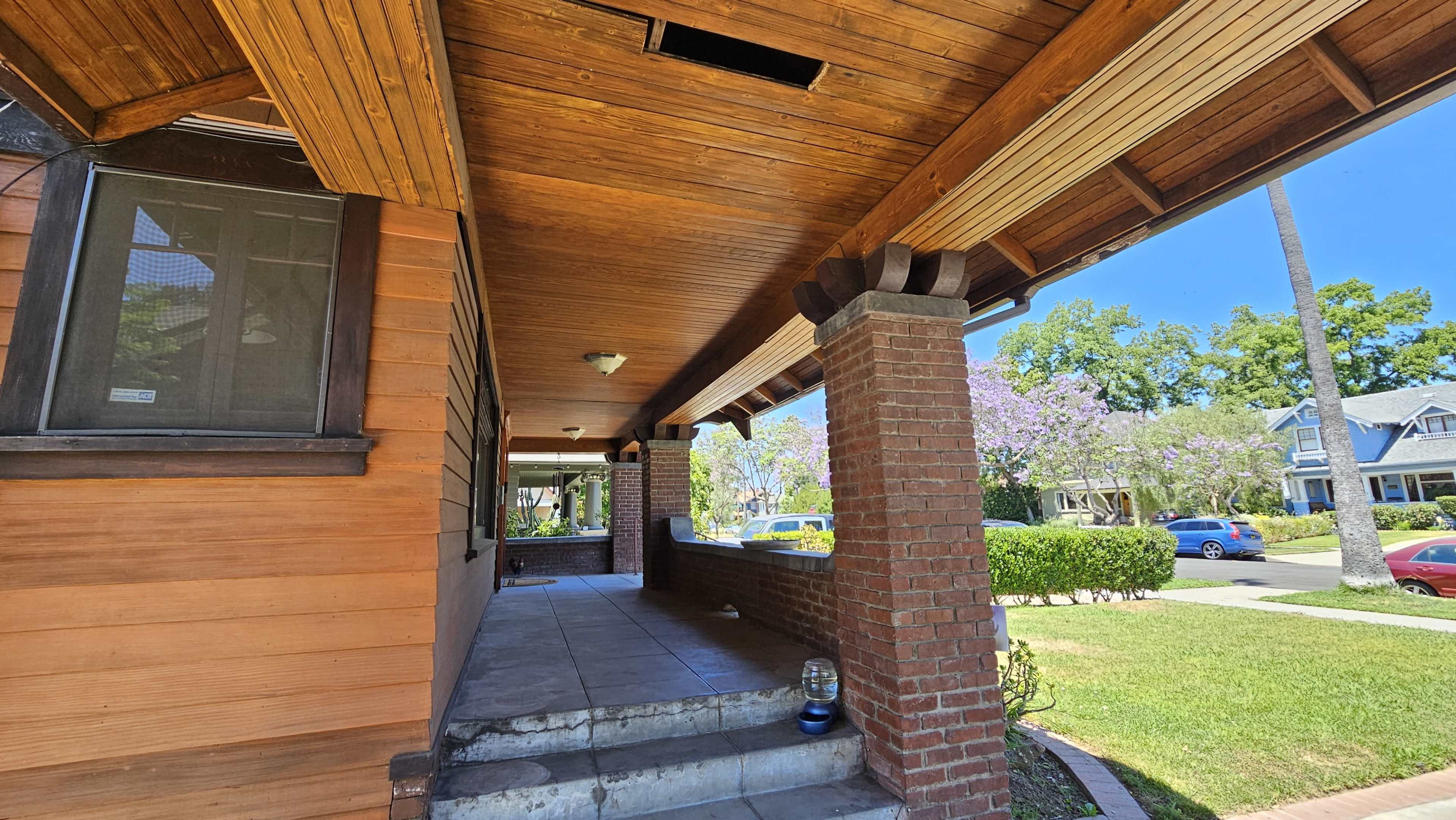 The image shows the entrance of a house with wooden walls, a tiled floor, and brick pillars, surrounded by greenery and purple flowering trees.