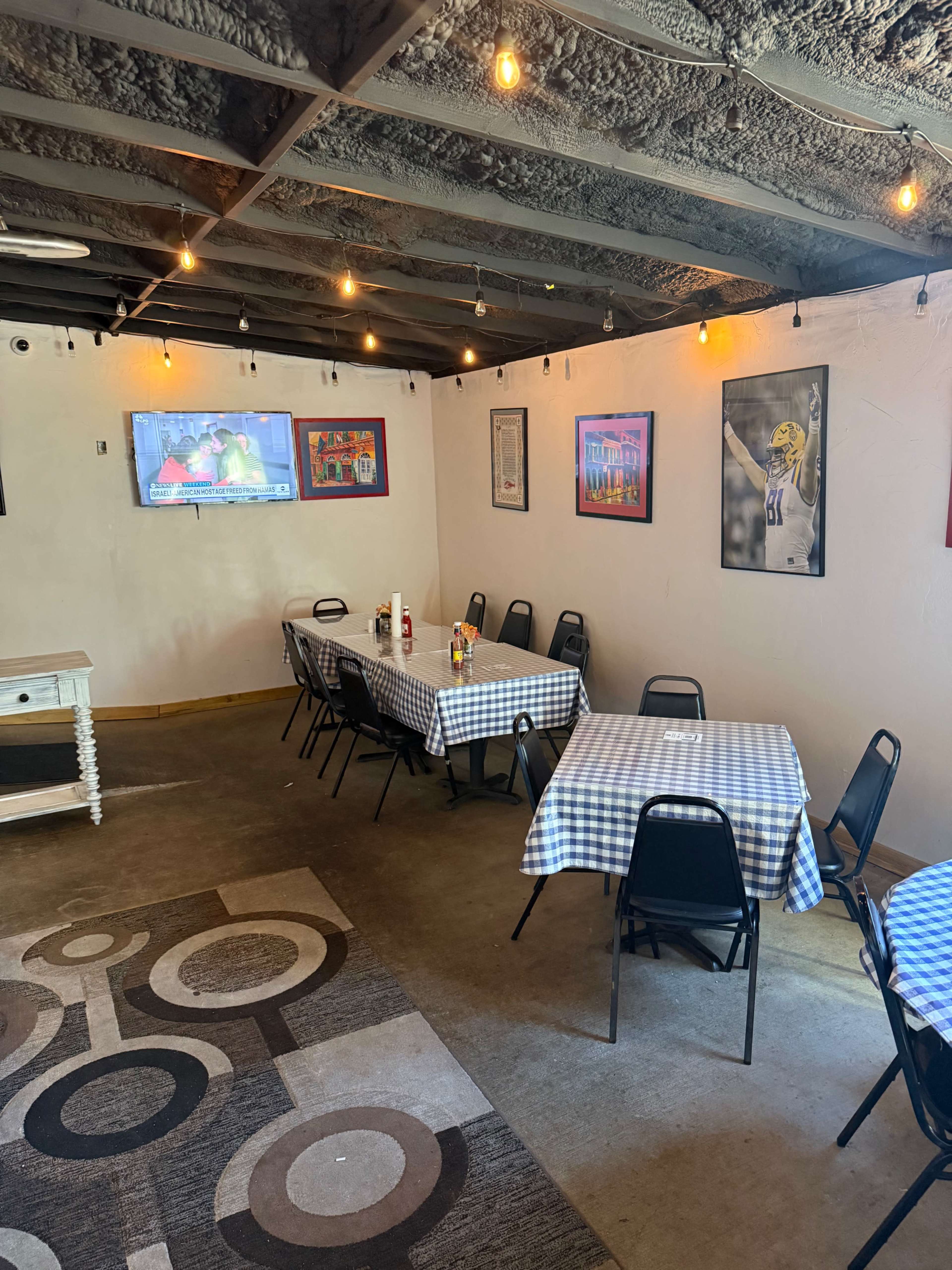 The image shows a dining area with checkered tablecloths on several tables, a TV mounted on the wall, and framed artwork hanging on the walls.