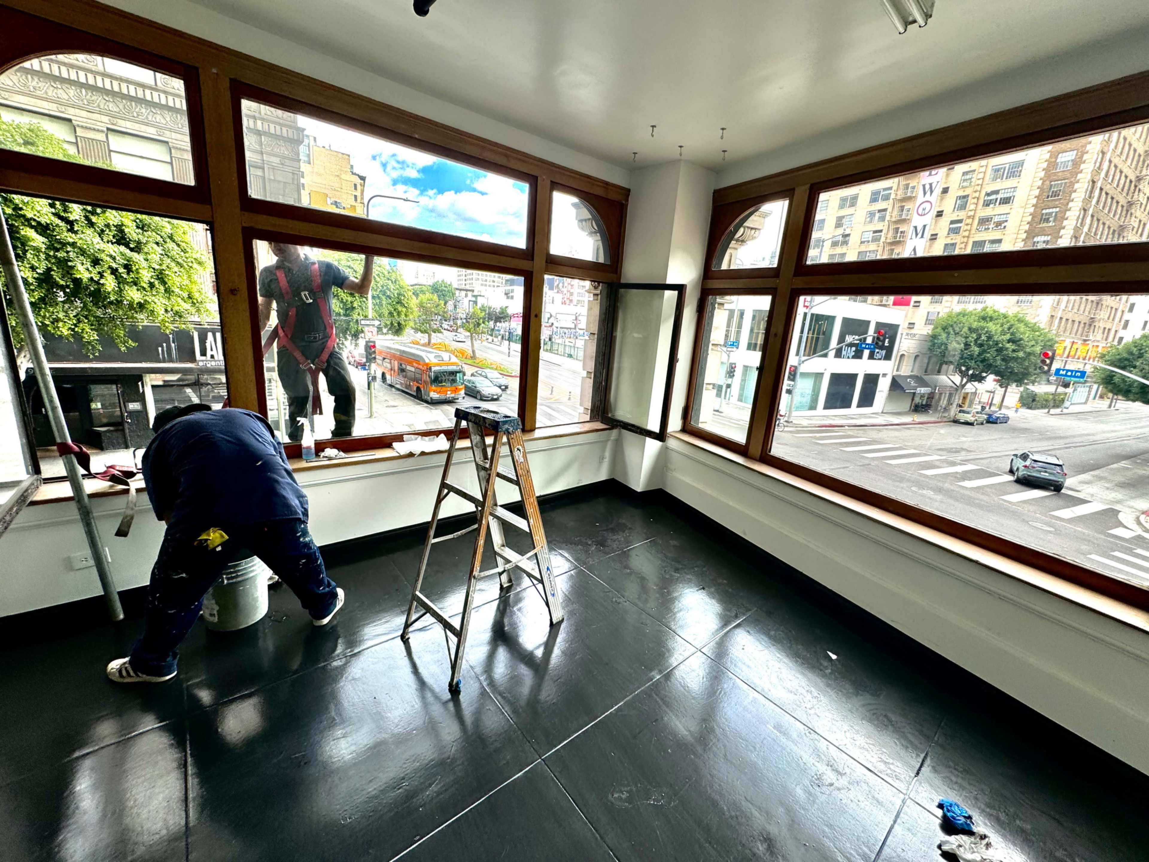 A person is painting the interior window frames of a vacant room with large windows overlooking a city street.