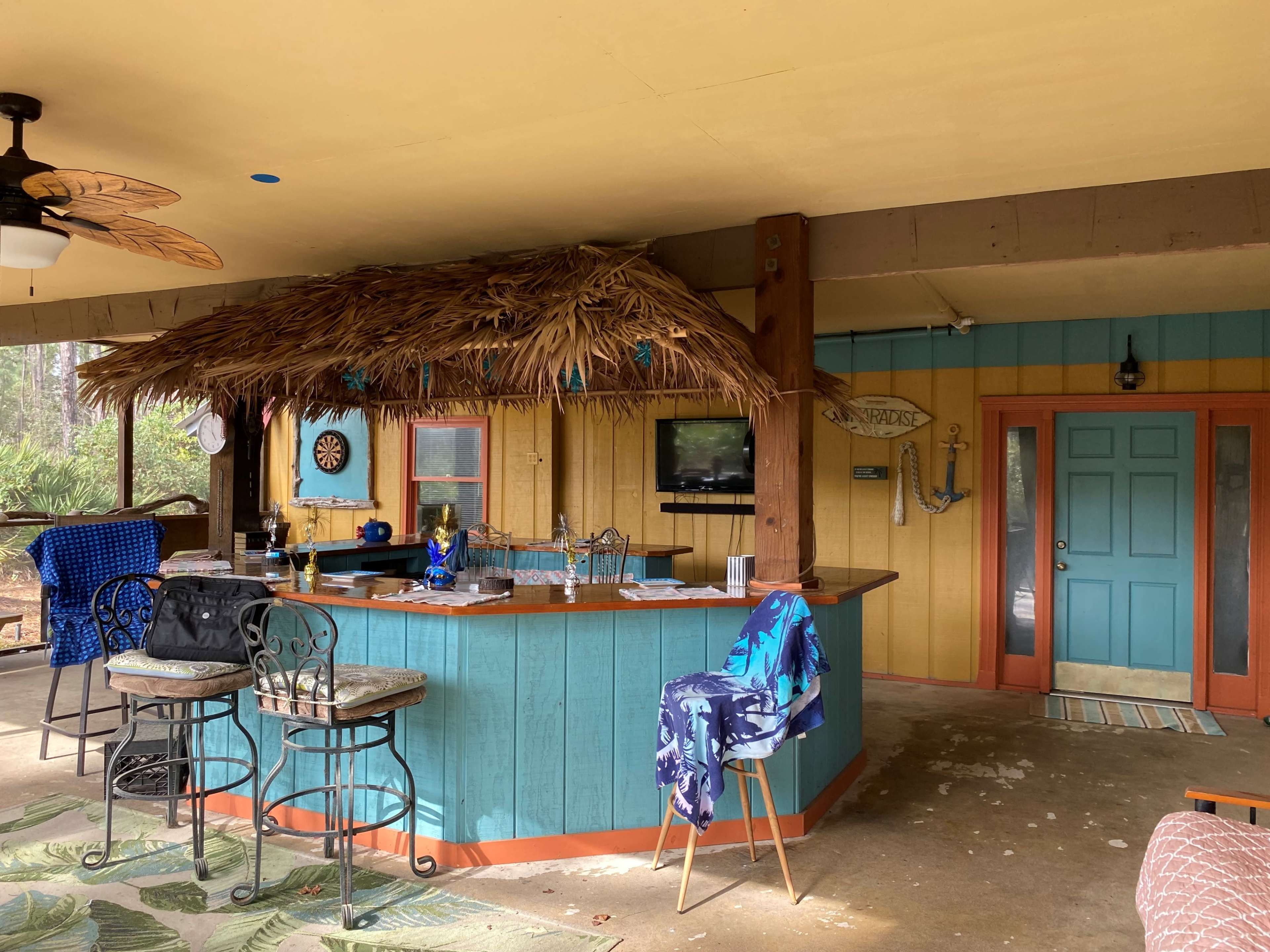 A covered outdoor bar area with a thatched roof, a blue bar counter, and a door leading to another room.