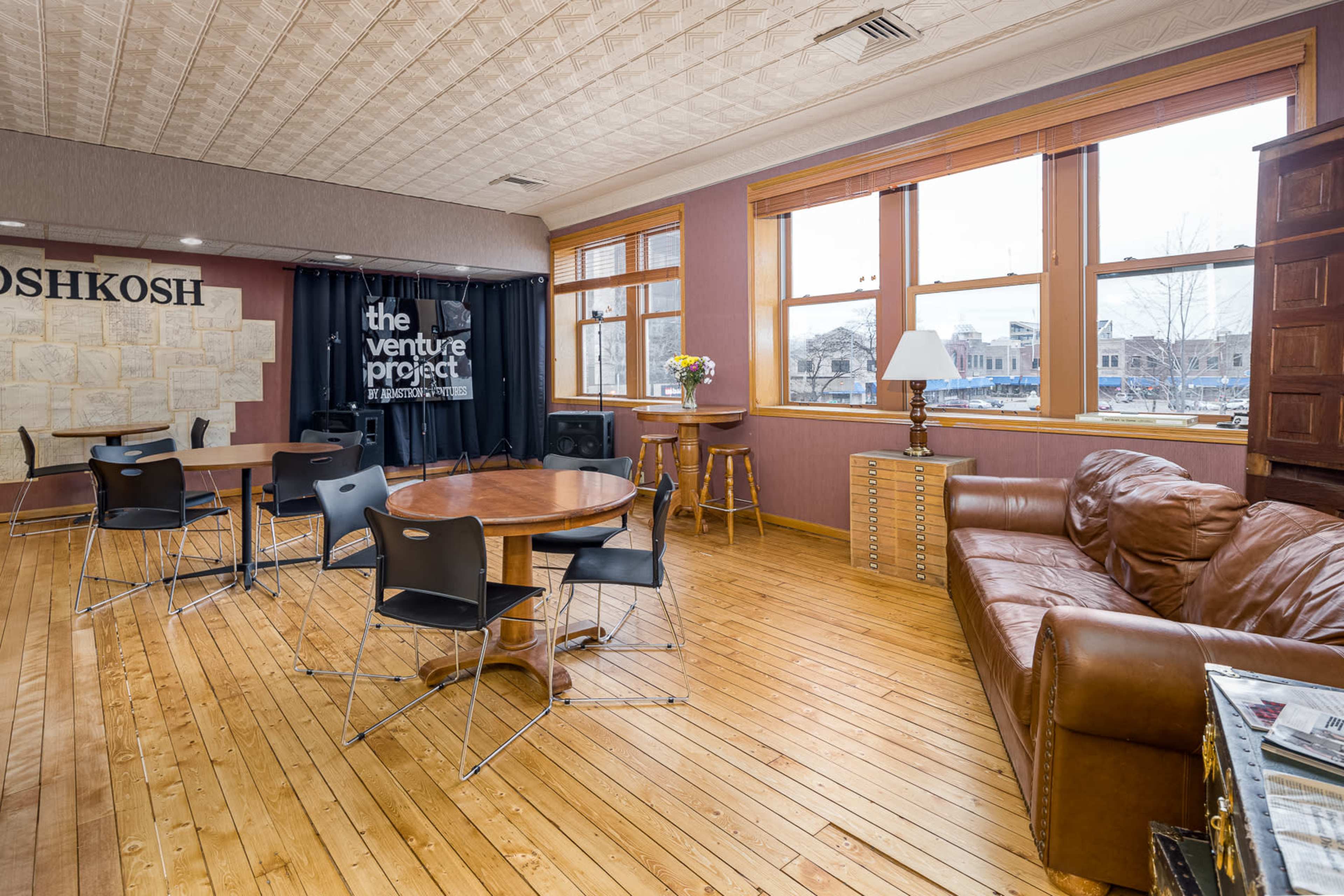 The room features wooden floors, large windows, a sofa, a round table with chairs, and a wall displaying a sign for "the venture project."