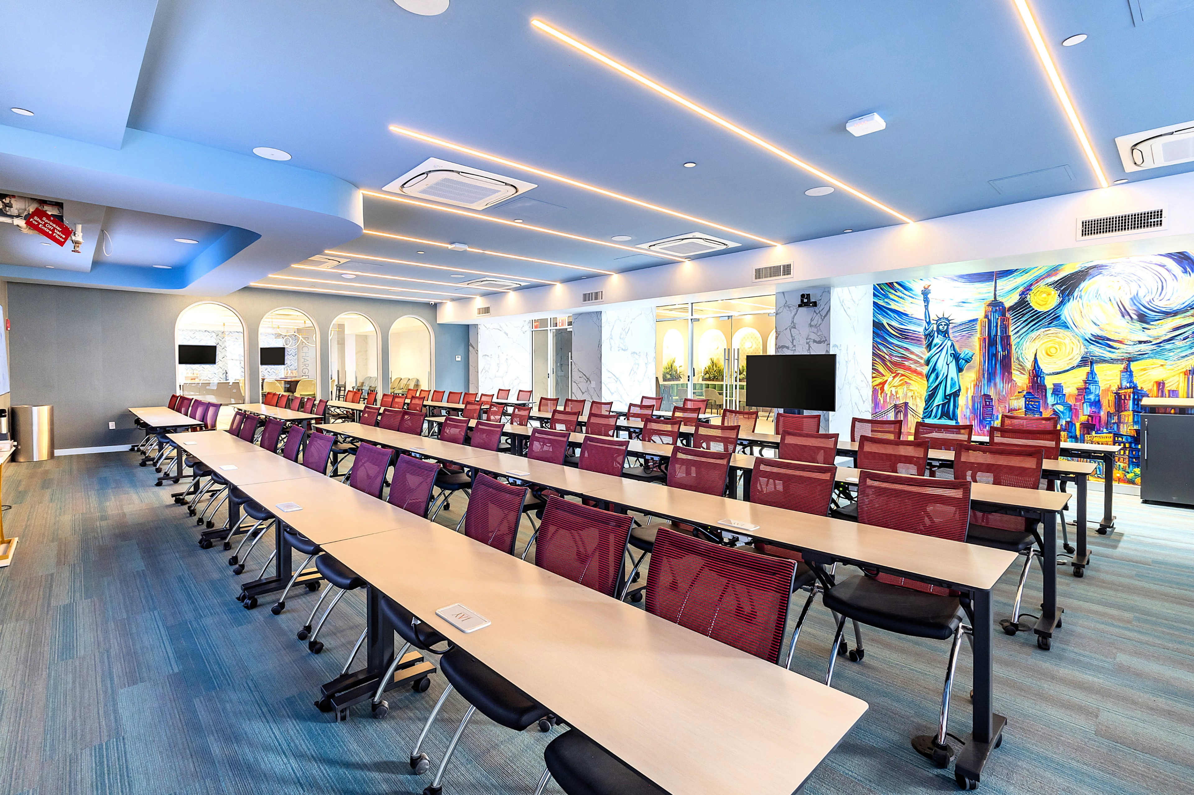The image shows a spacious conference room with long tables arranged in rows, equipped with purple chairs, and featuring modern decor including a mural on one wall.