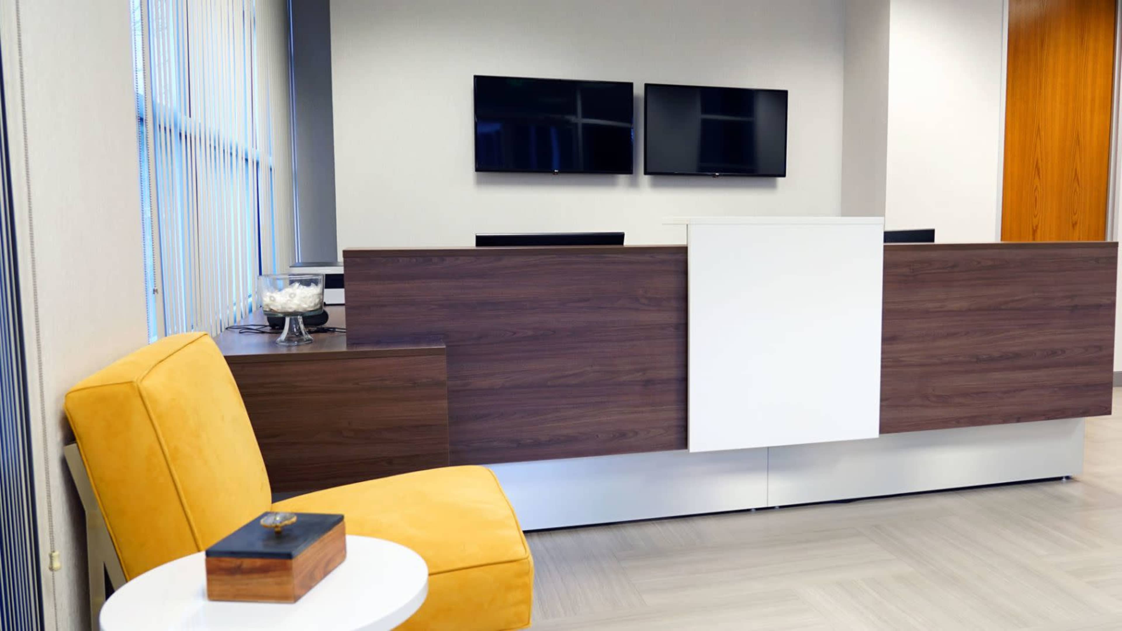 The image shows a reception area featuring a wooden front desk, two mounted televisions, a yellow armchair, and a small white table.