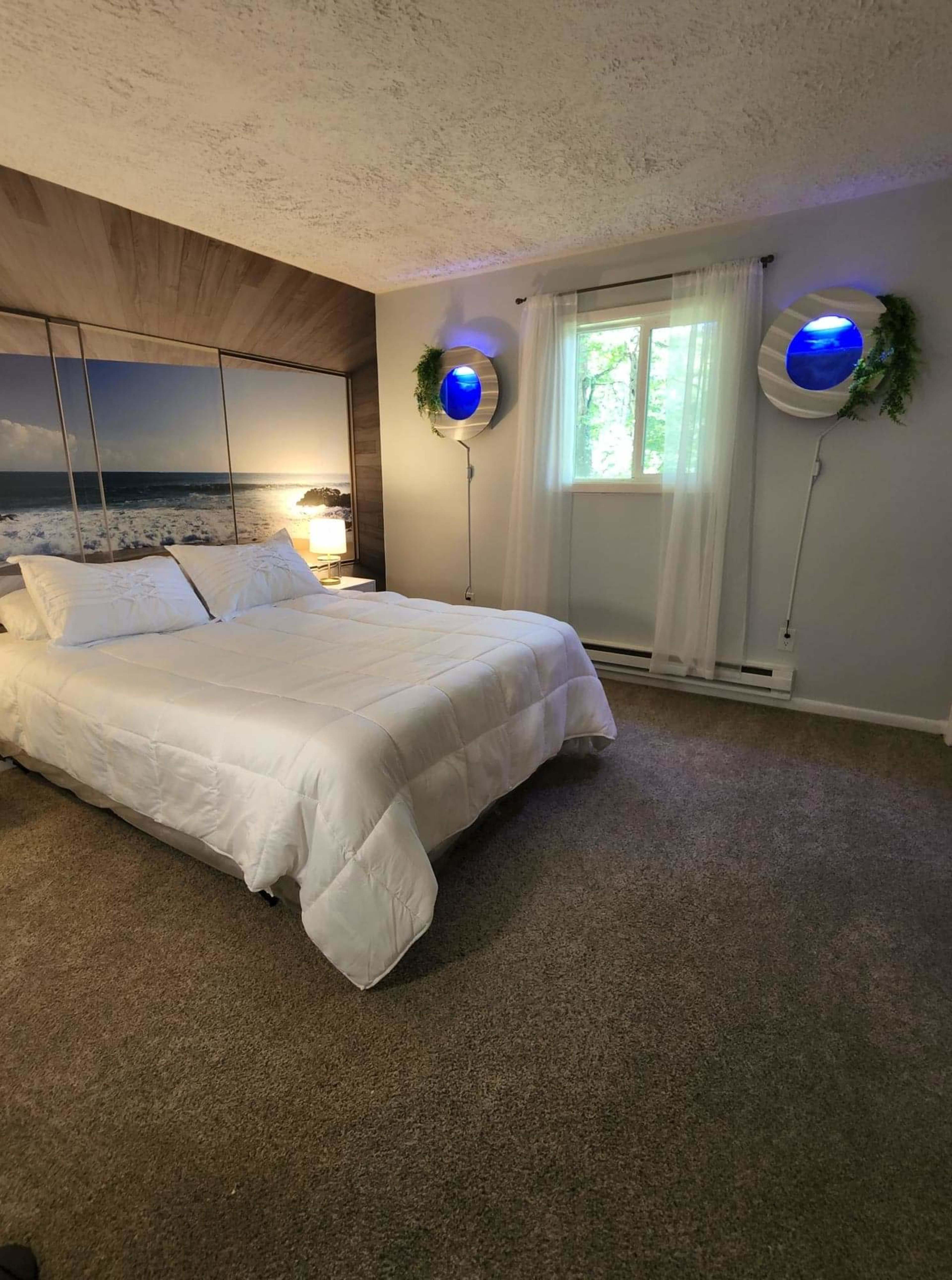 A bedroom features a bed with a white comforter, a large photo of a beach on the wall, and two round wall-mounted lights with blue accents.