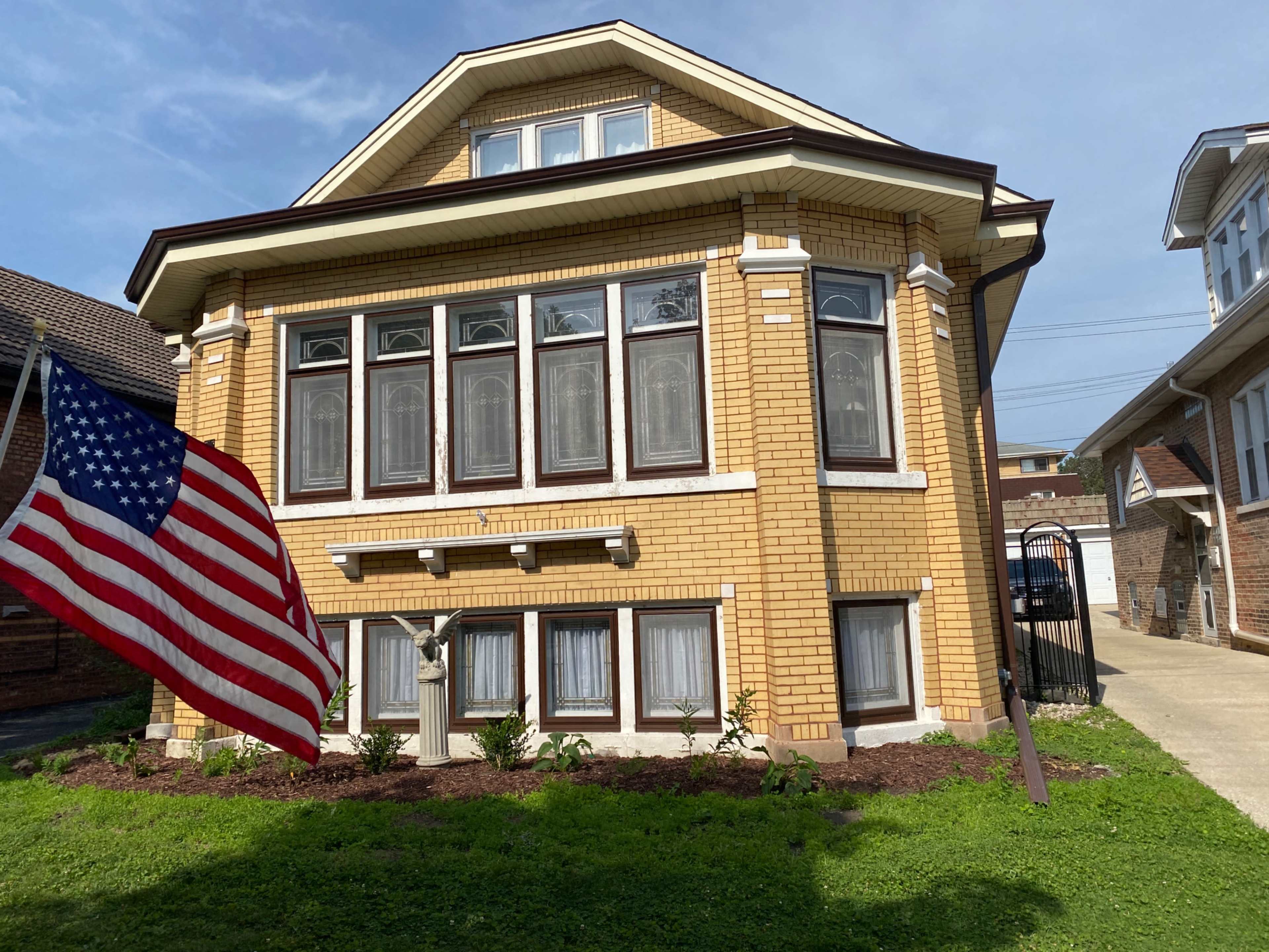 A brick house with multiple large windows features an American flag prominently displayed in the front yard.