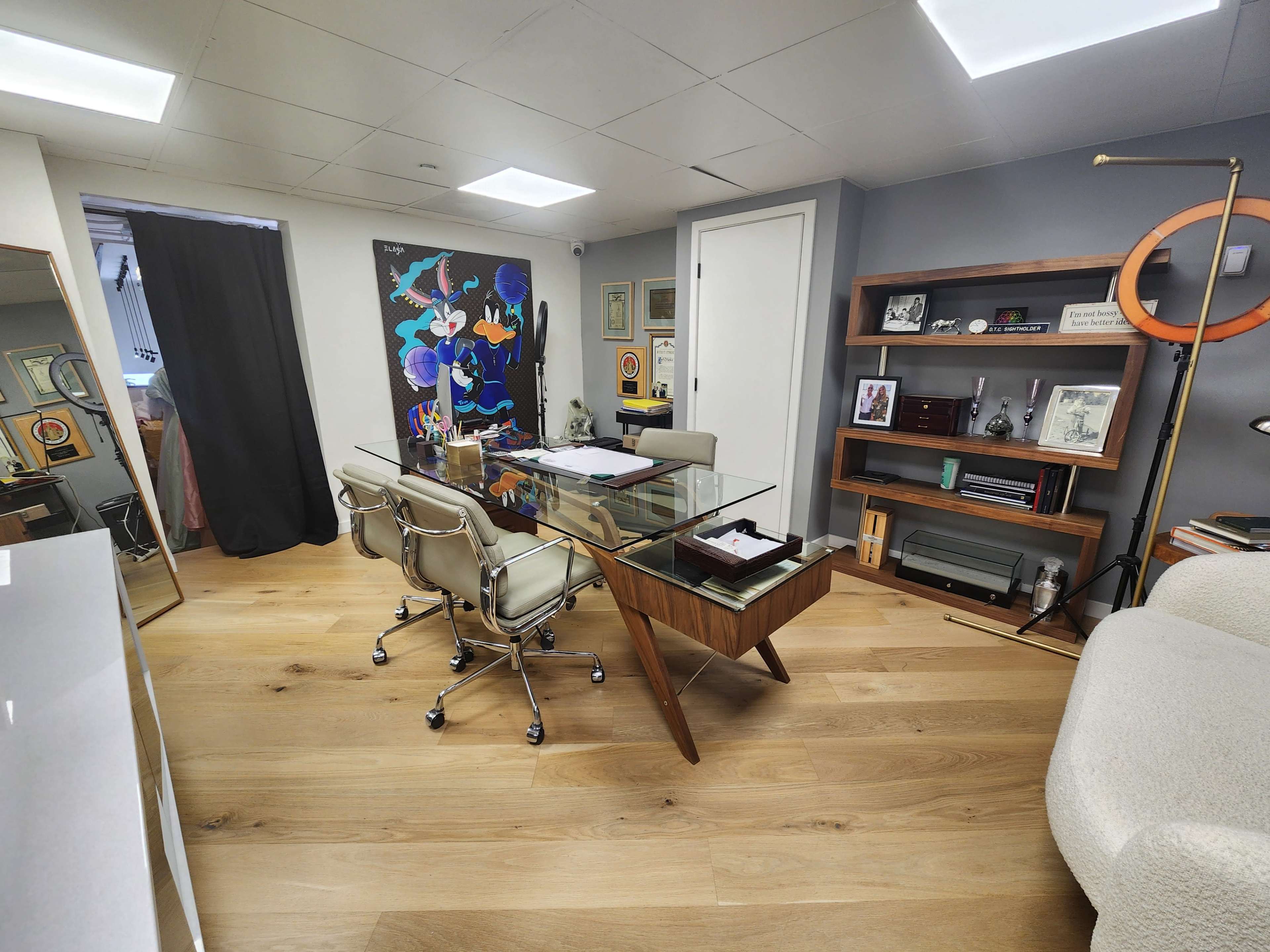 The image shows a modern office space featuring a glass-top desk with three office chairs, a bookshelf, and artwork on the wall.