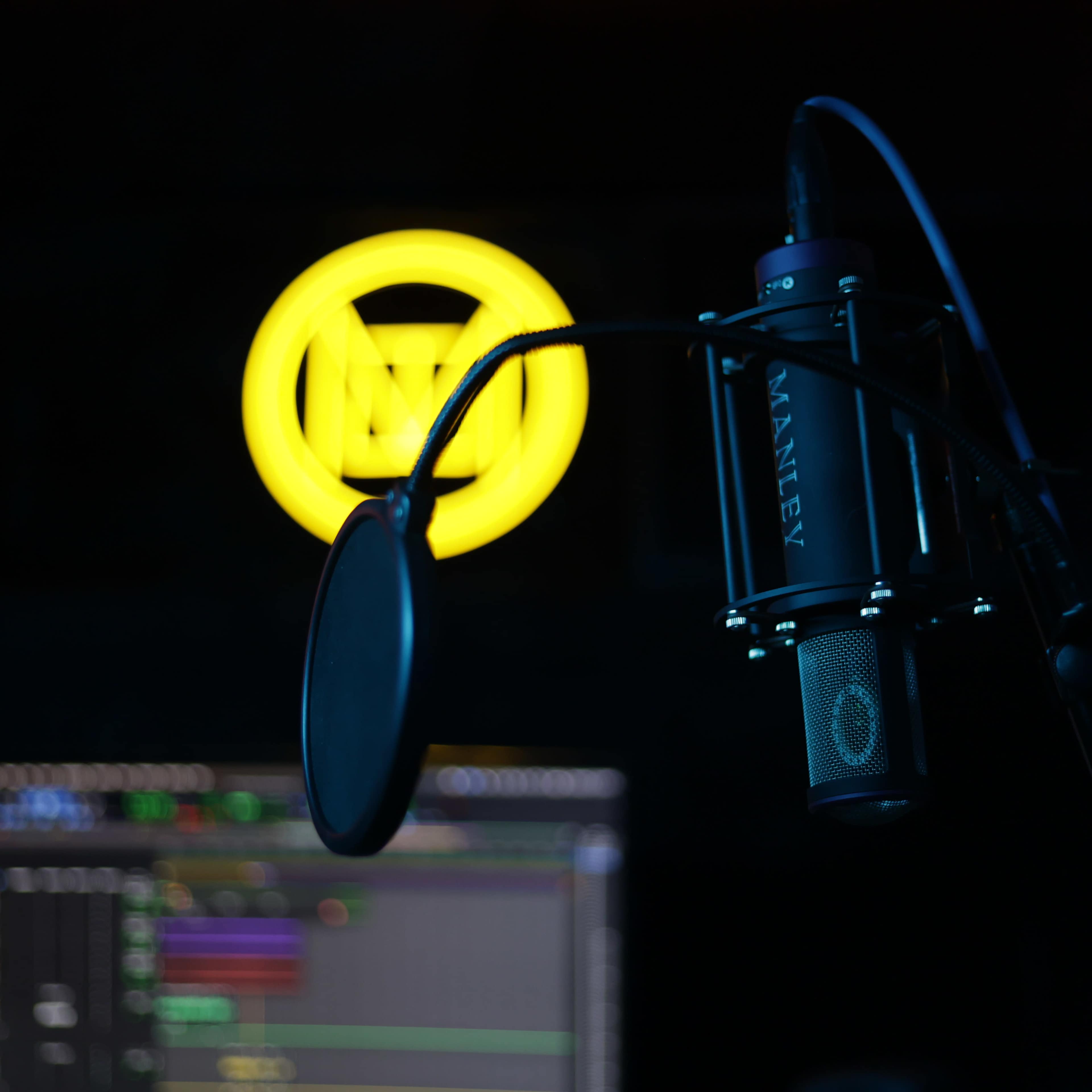 A microphone with a pop filter is positioned in front of a computer screen displaying audio editing software, with a yellow circular logo illuminated in the background.