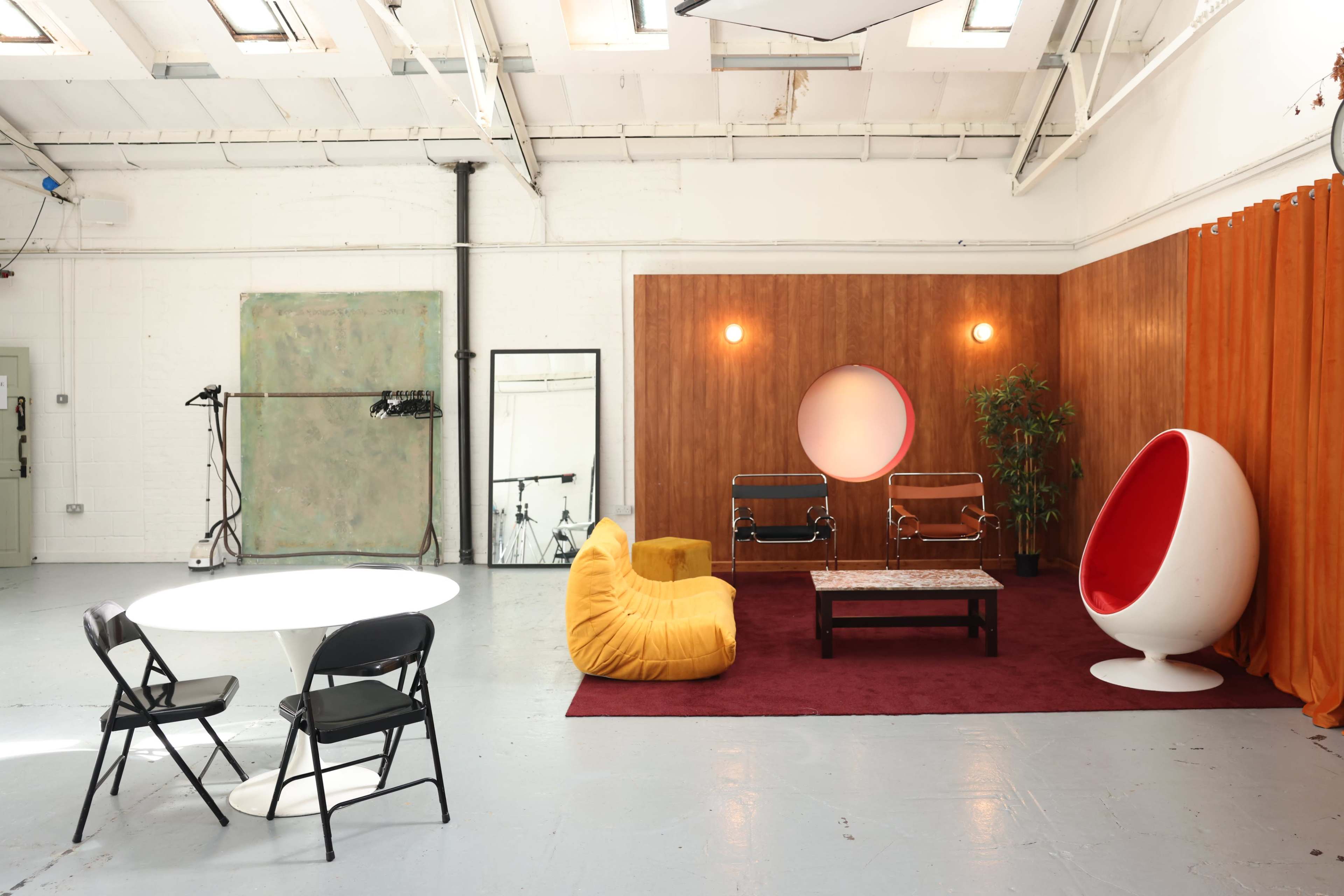 The scene features a modern studio space with a yellow beanbag chair, a red egg-shaped chair, a circular mirror, and a white dining table surrounded by black folding chairs on a maroon rug.