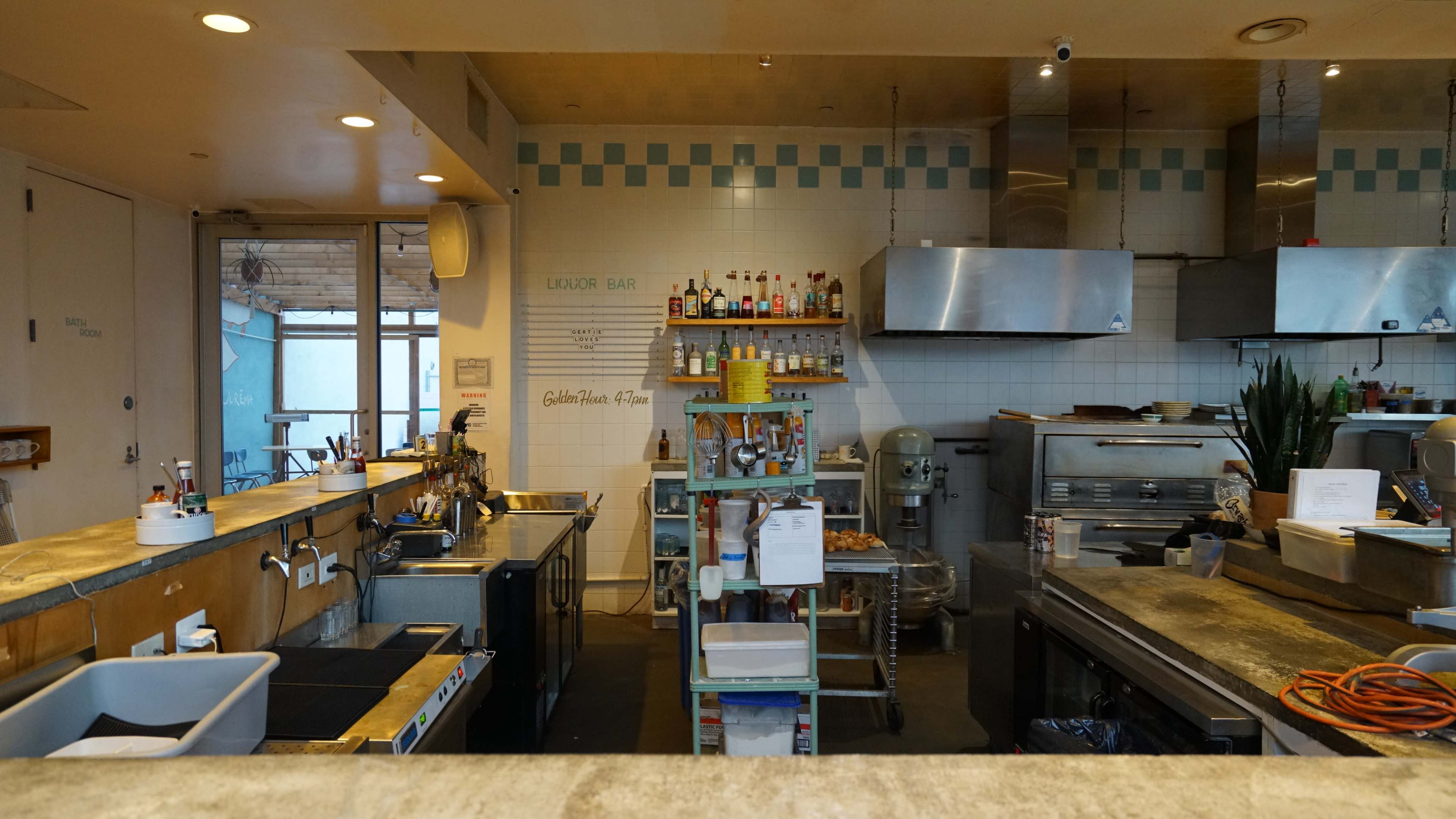 The image shows a kitchen with stainless steel appliances, shelves stocked with bottles and ingredients, and a counter area with food preparation equipment.