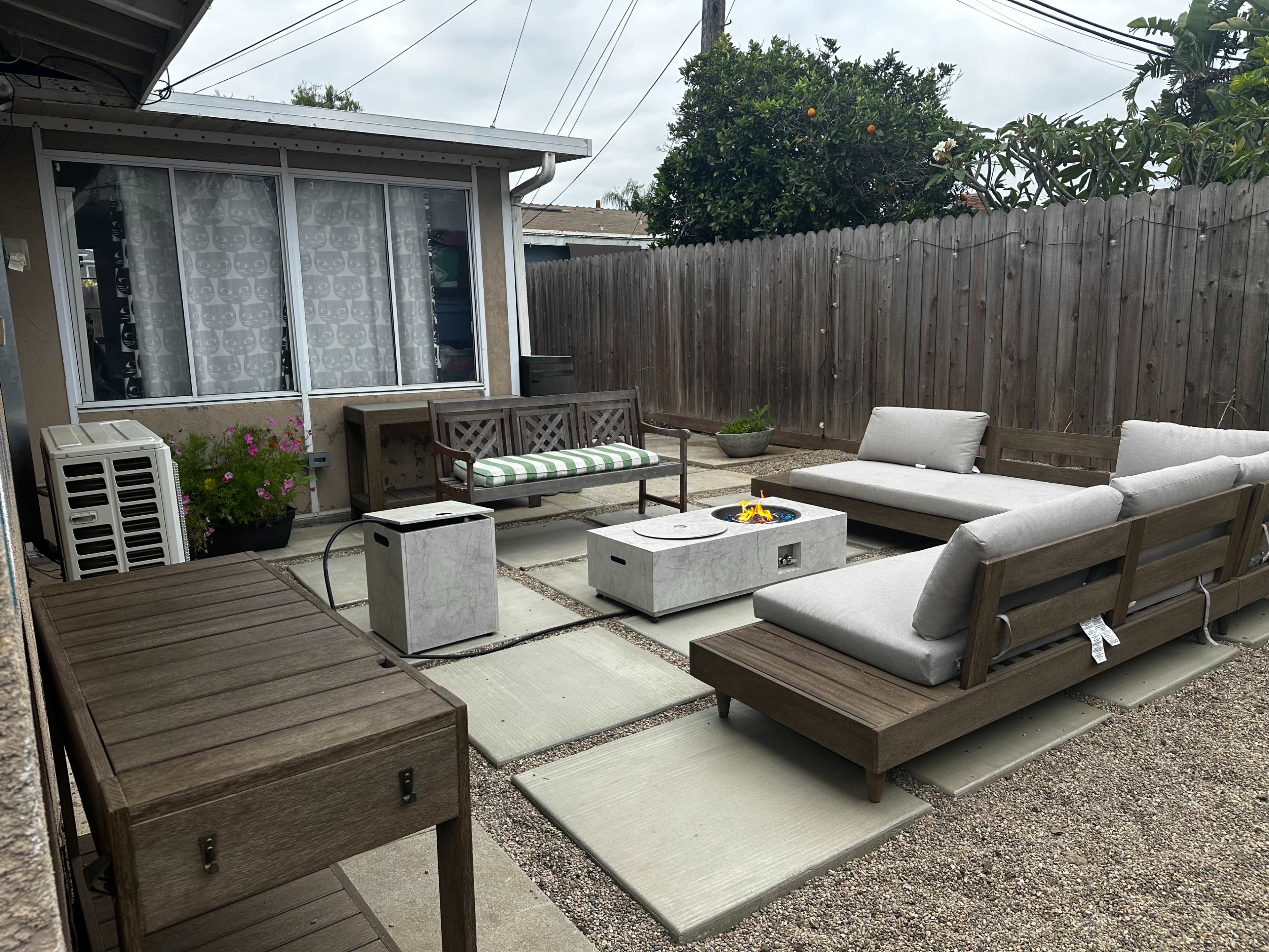 The image shows a modern outdoor living space with gray seating, a fire pit, decorative plants, and a wooden fence in the background.