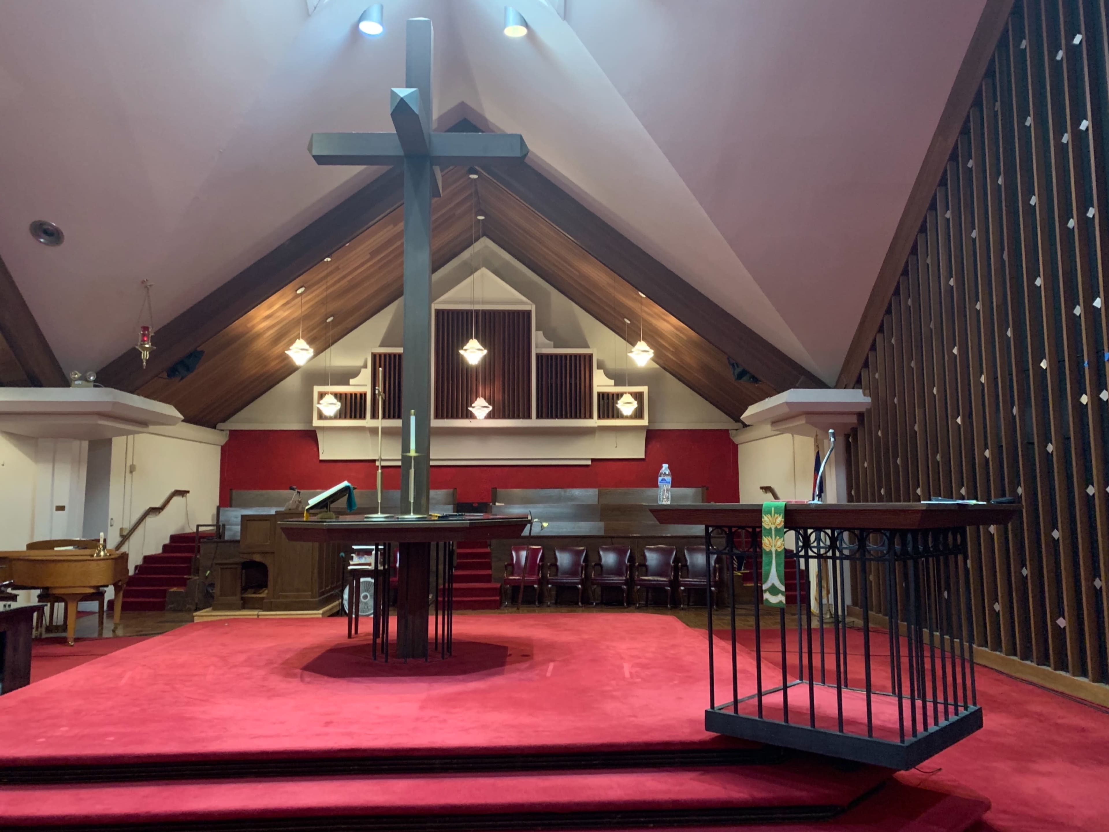 The interior of a church features a large cross at the center of a raised altar area, surrounded by pews and decorative elements against a backdrop of wooden architecture.