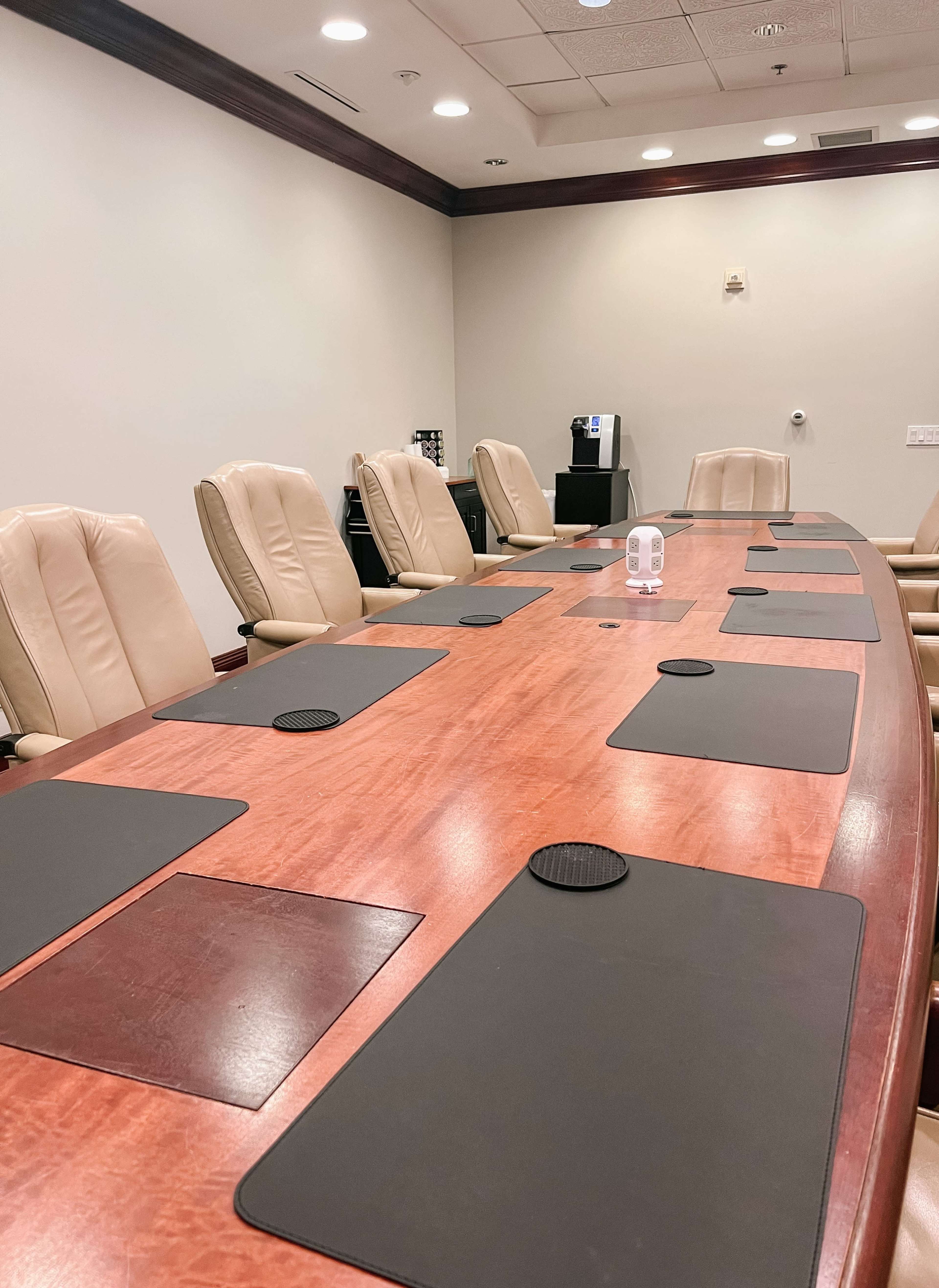 A long wooden conference table with beige chairs and black placemats is set up in a well-lit meeting room.