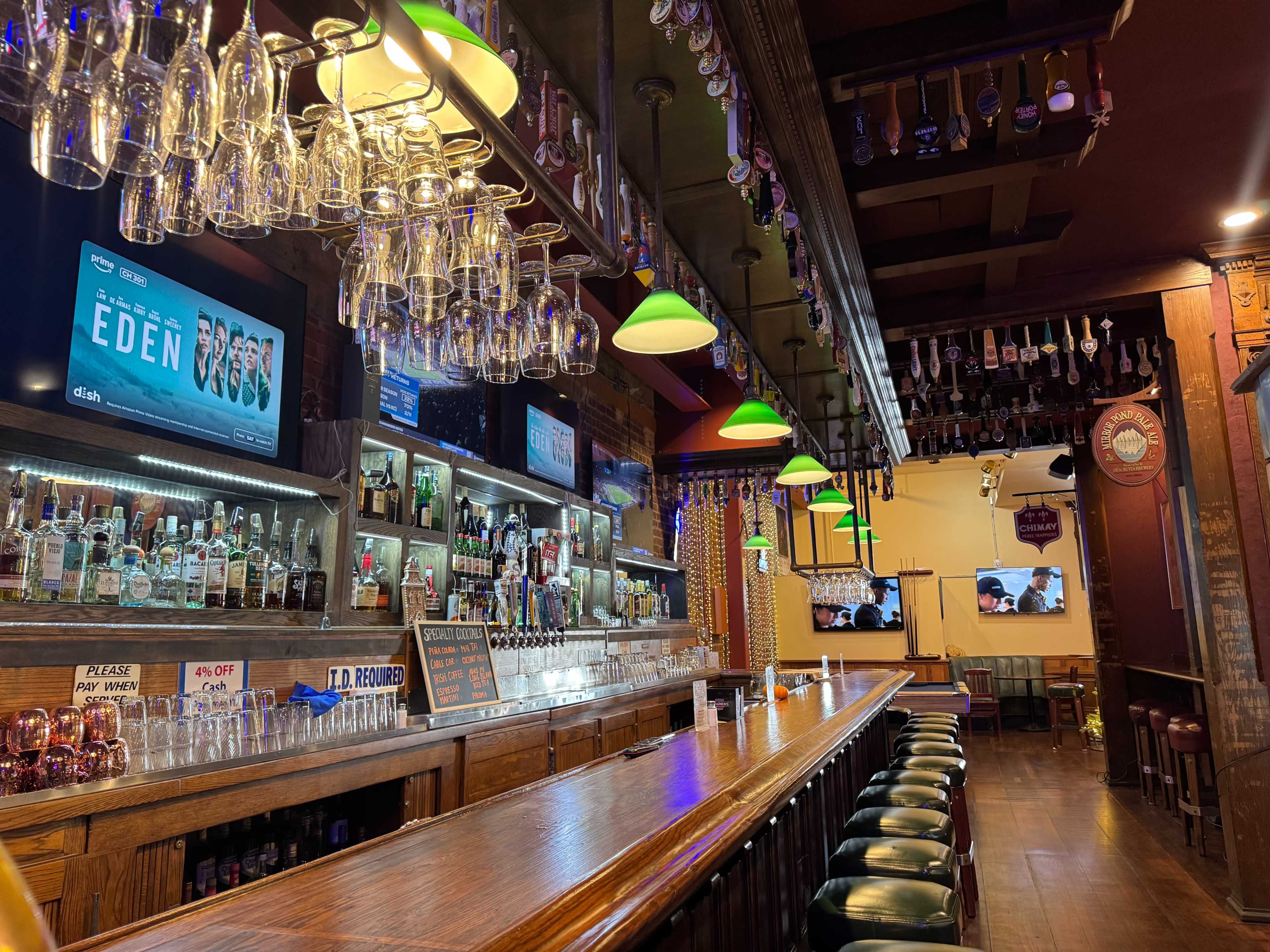 The image shows a well-lit bar with a wooden counter, shelves lined with various bottles, and two screens displaying content.