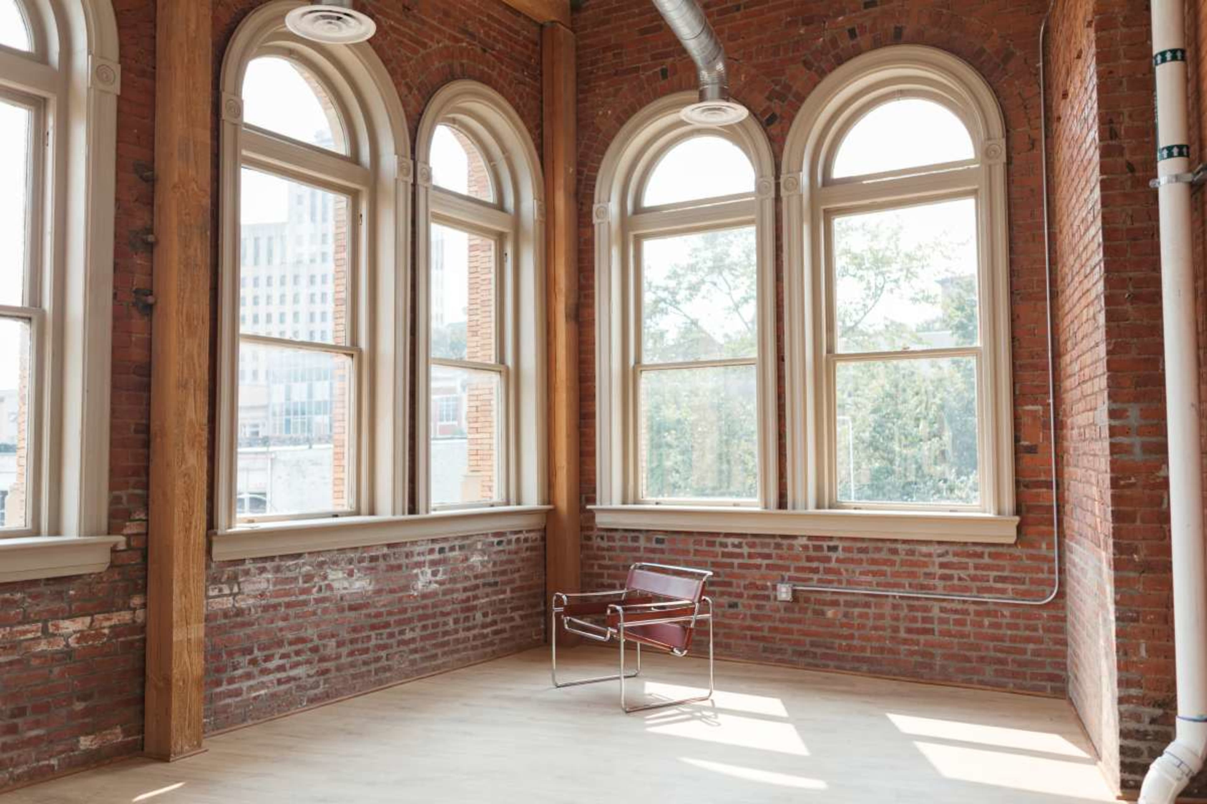 The image shows a vacant room with large arched windows, exposed brick walls, and a single chair positioned in the center.