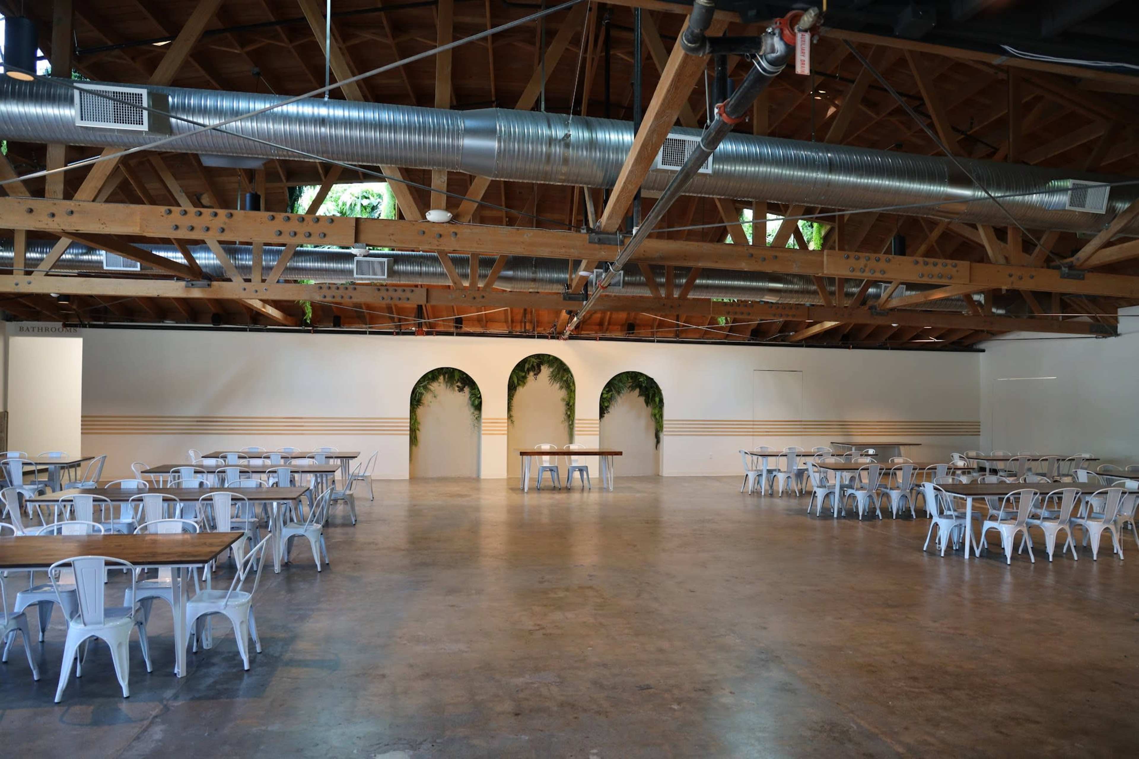 The image shows a spacious event hall with wooden beams, metal tables and chairs, and decorative arches on the walls.