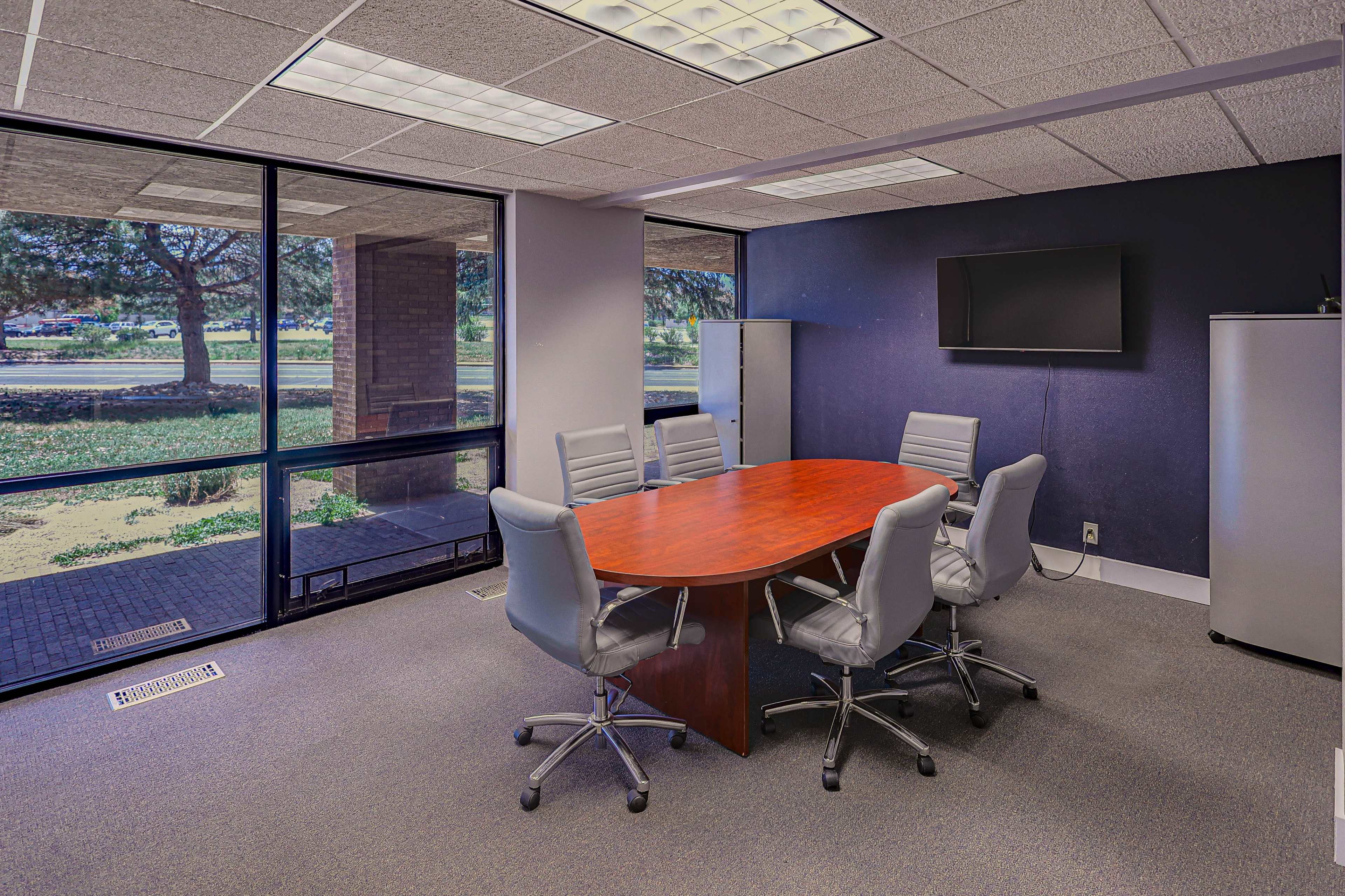 The image shows a conference room with a round wooden table surrounded by four gray chairs and a television mounted on the wall.