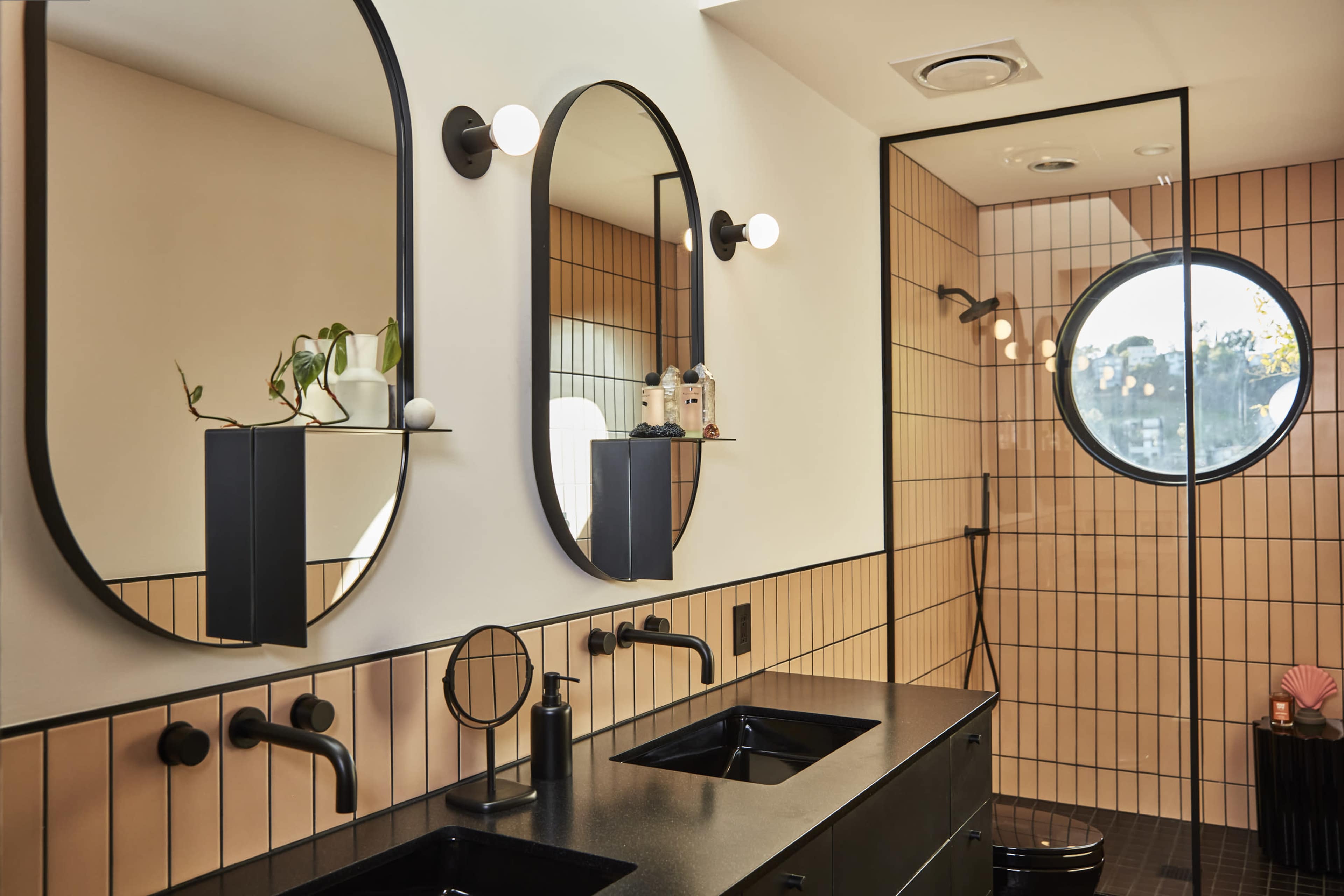 The bathroom features two black oval mirrors, a double sink with a dark countertop, and a glass shower enclosed by modern tile walls in shades of peach and orange.