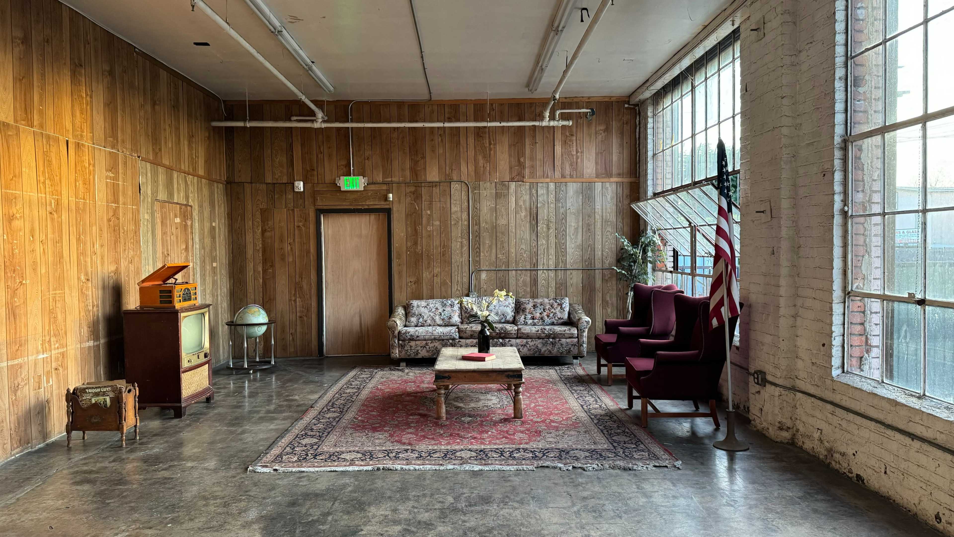 A spacious interior features a wooden paneled wall, a floral-patterned sofa, two red chairs, a coffee table, and a vintage television set beside a globe.