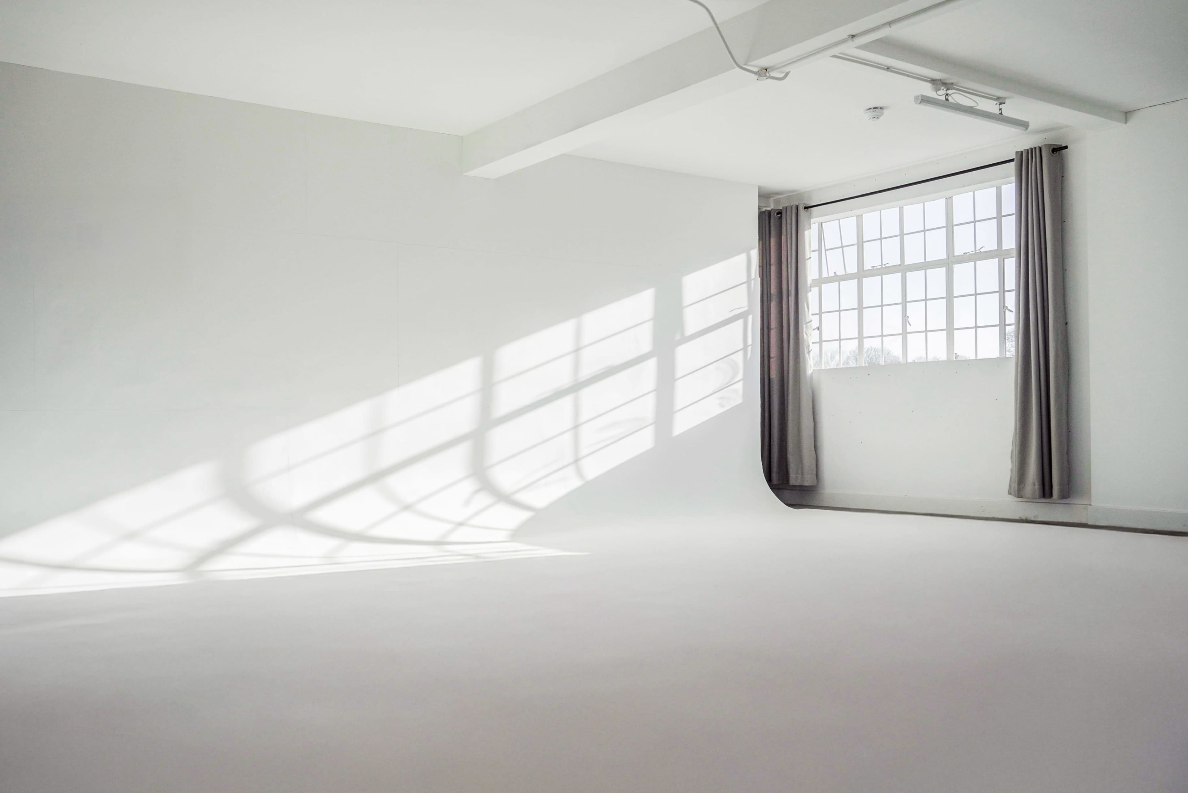 The image shows a bright, minimalist room with white walls, a large window, and soft shadows cast across the floor.