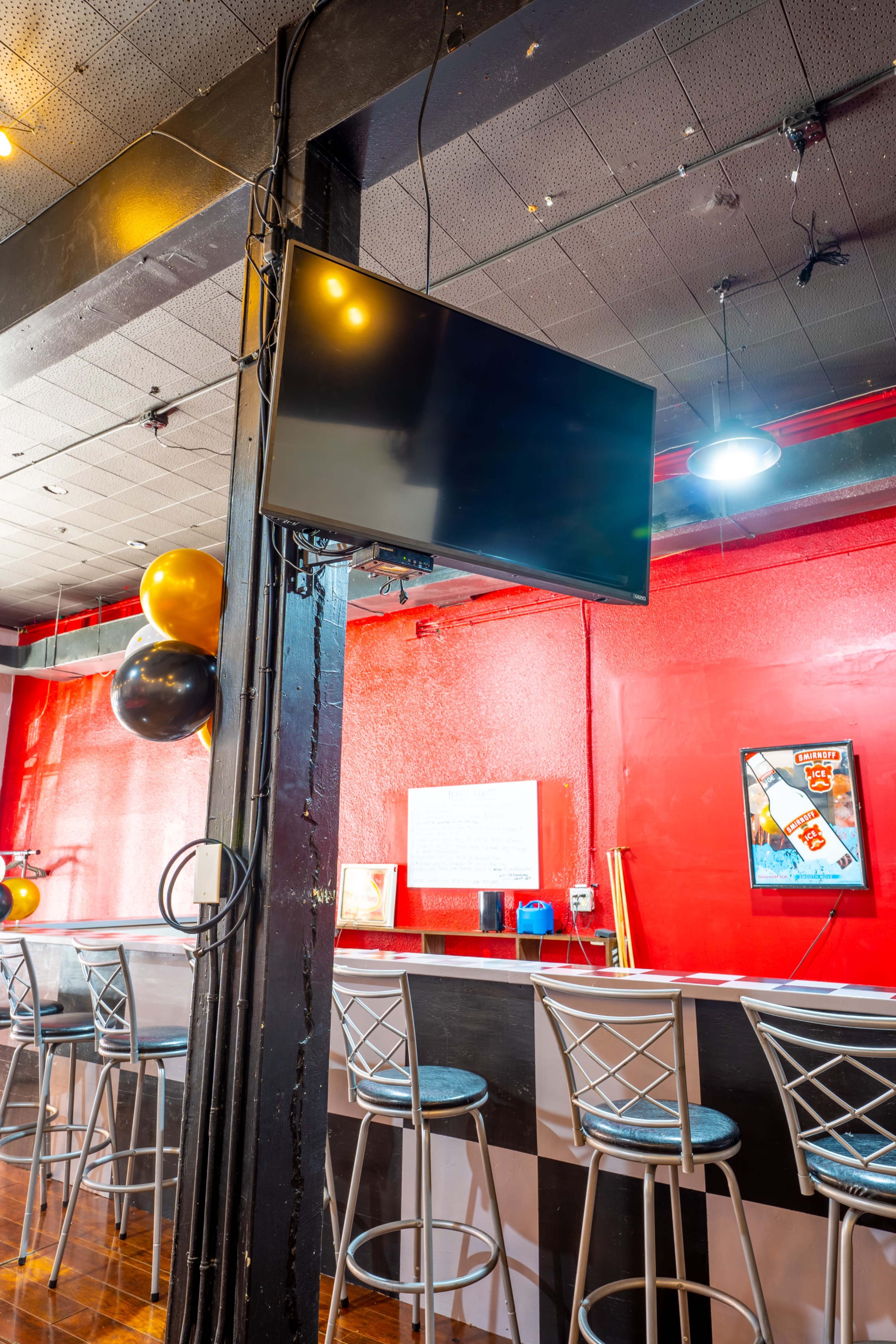 The image shows a bar area with a wall-mounted television, red walls, and several metal stools arranged along a counter.