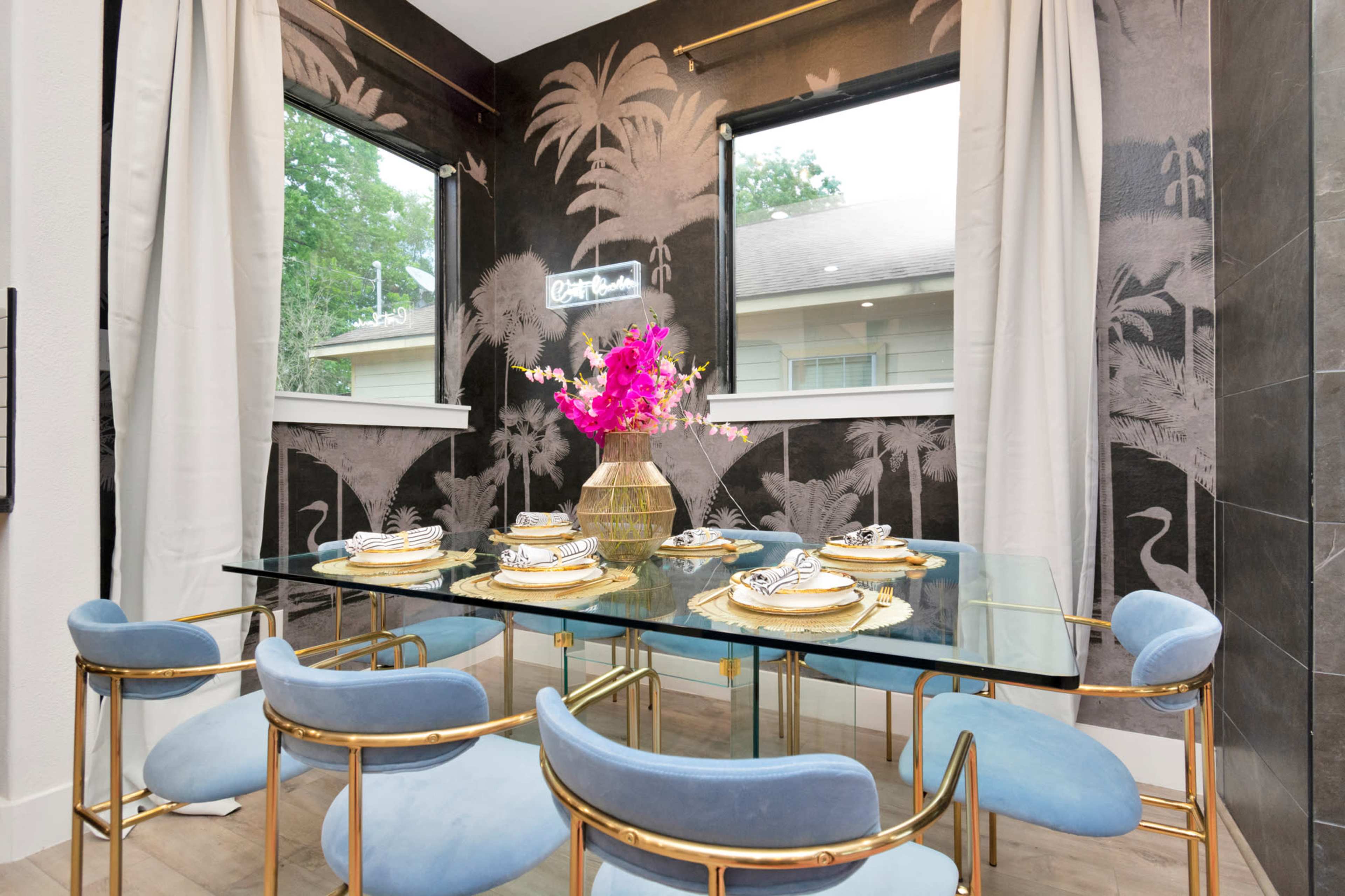 A dining area features a glass table set with dishes and gold accents, surrounded by blue chairs and adorned with pink flowers against a patterned wall.