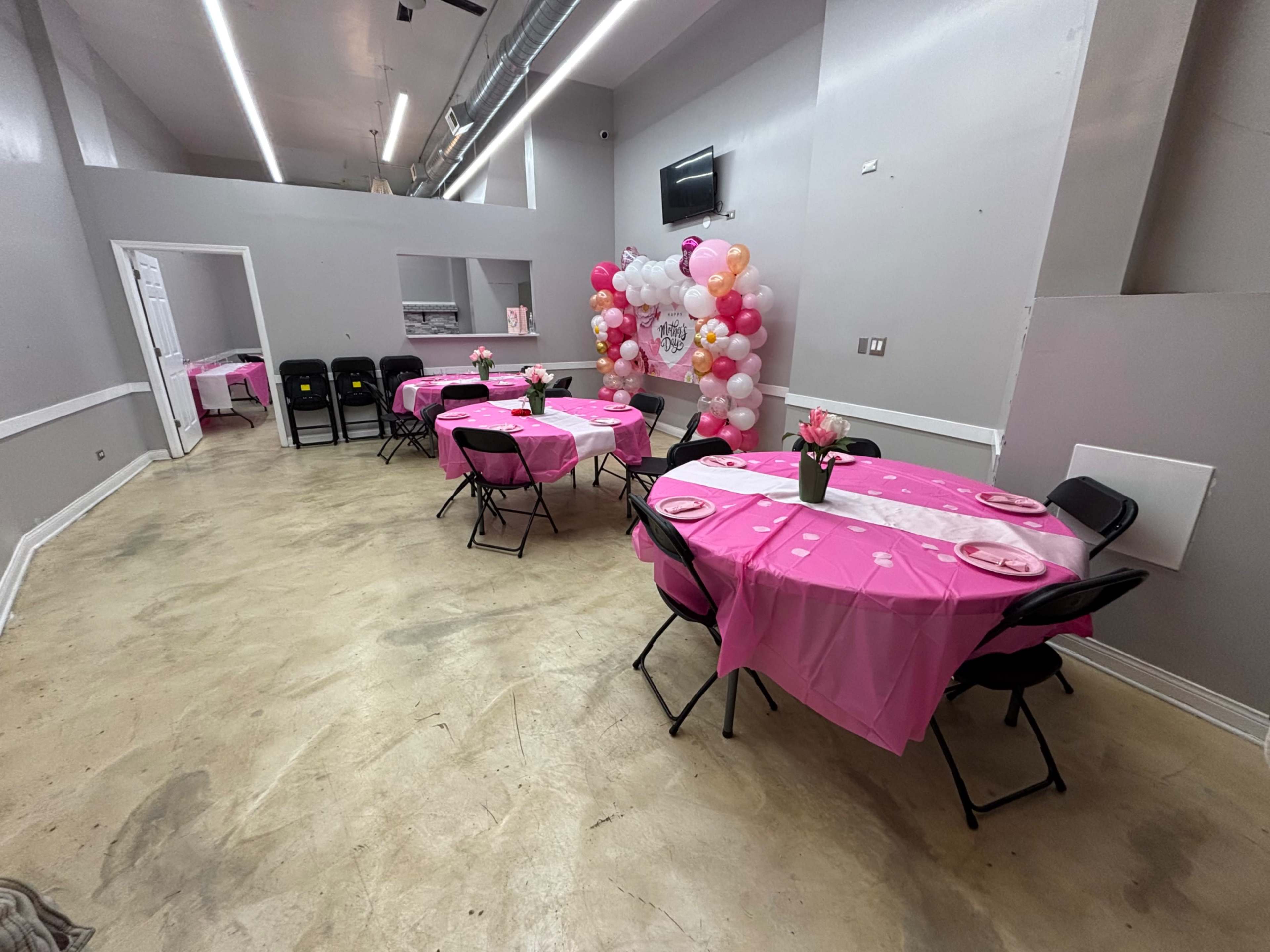 The image shows a decorated event space with round tables covered in pink tablecloths, black chairs, and a balloon arch at one end.