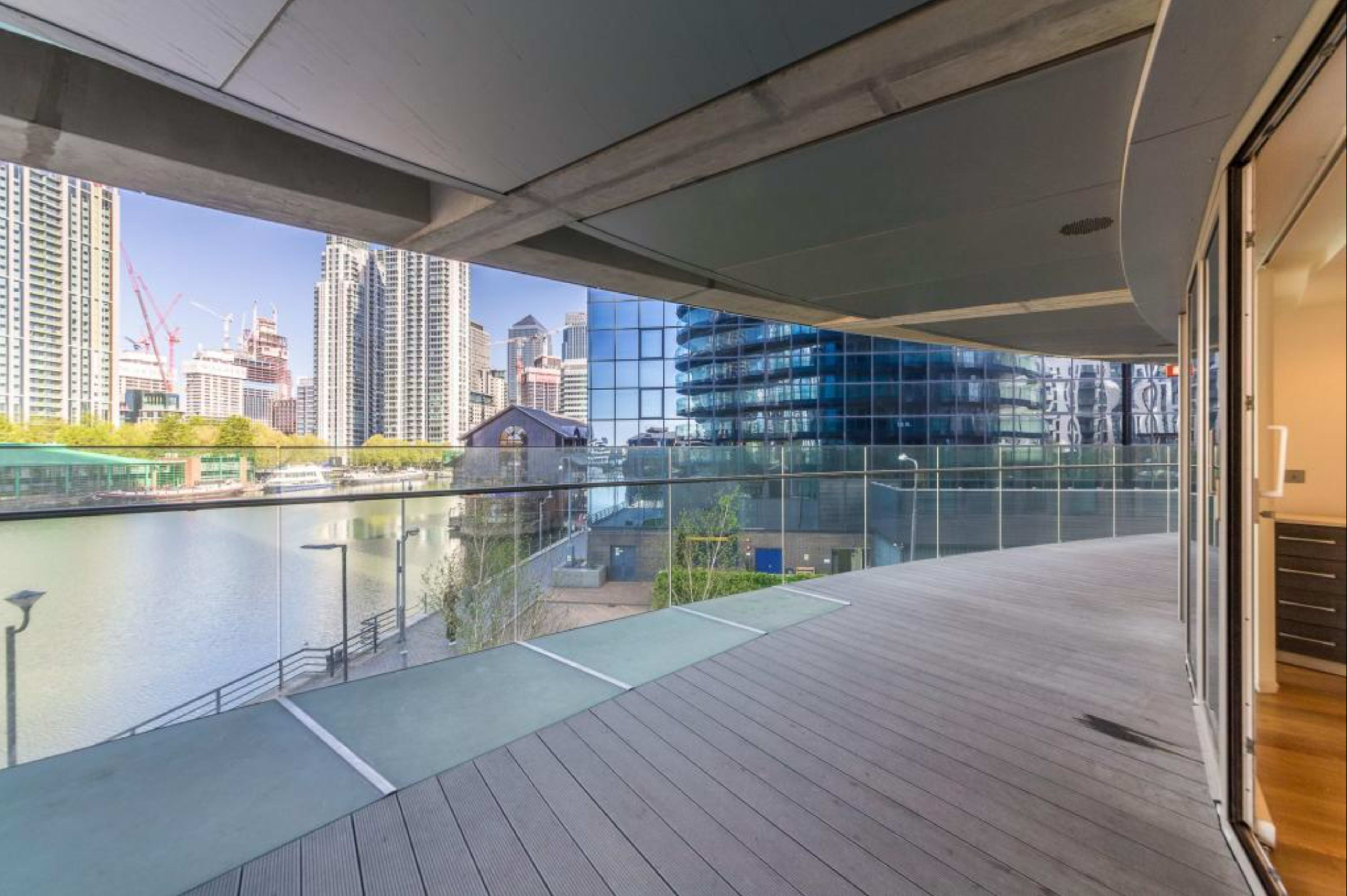 The image shows a spacious balcony with a wooden deck overlooking a body of water and modern buildings in an urban setting.