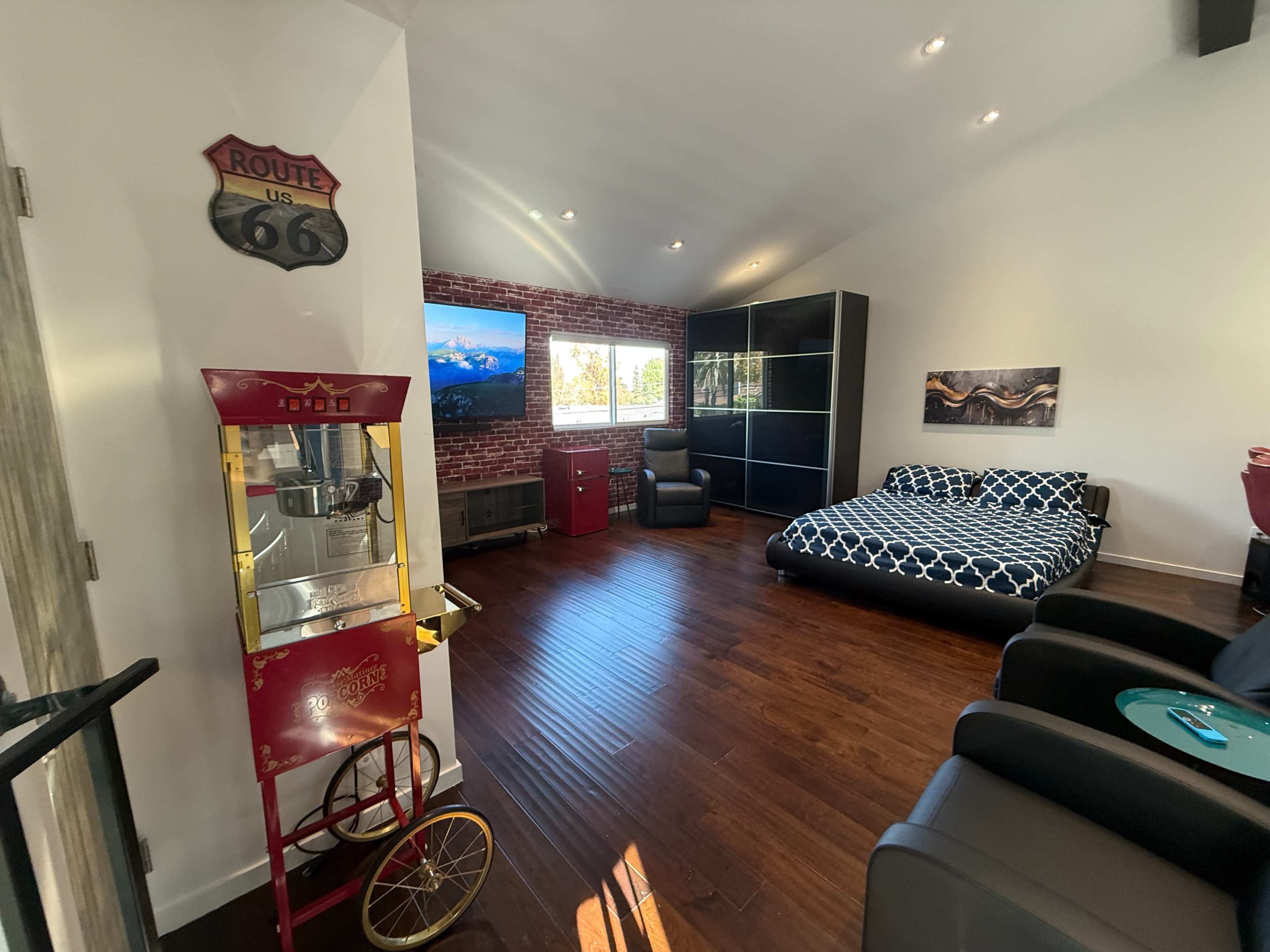 The room features a bed with a patterned comforter, two black lounge chairs, a vintage popcorn machine, and a large window with natural light illuminating the wooden floor.