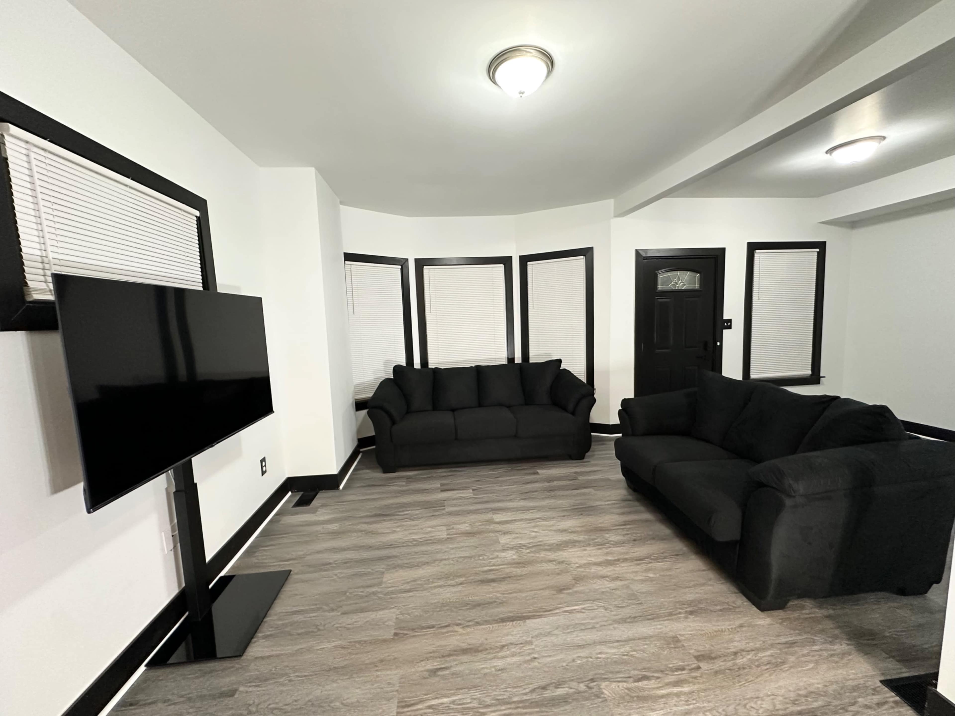 The image shows a living room with two black sofas facing a television mounted on a stand, featuring windows with white blinds and a black door.