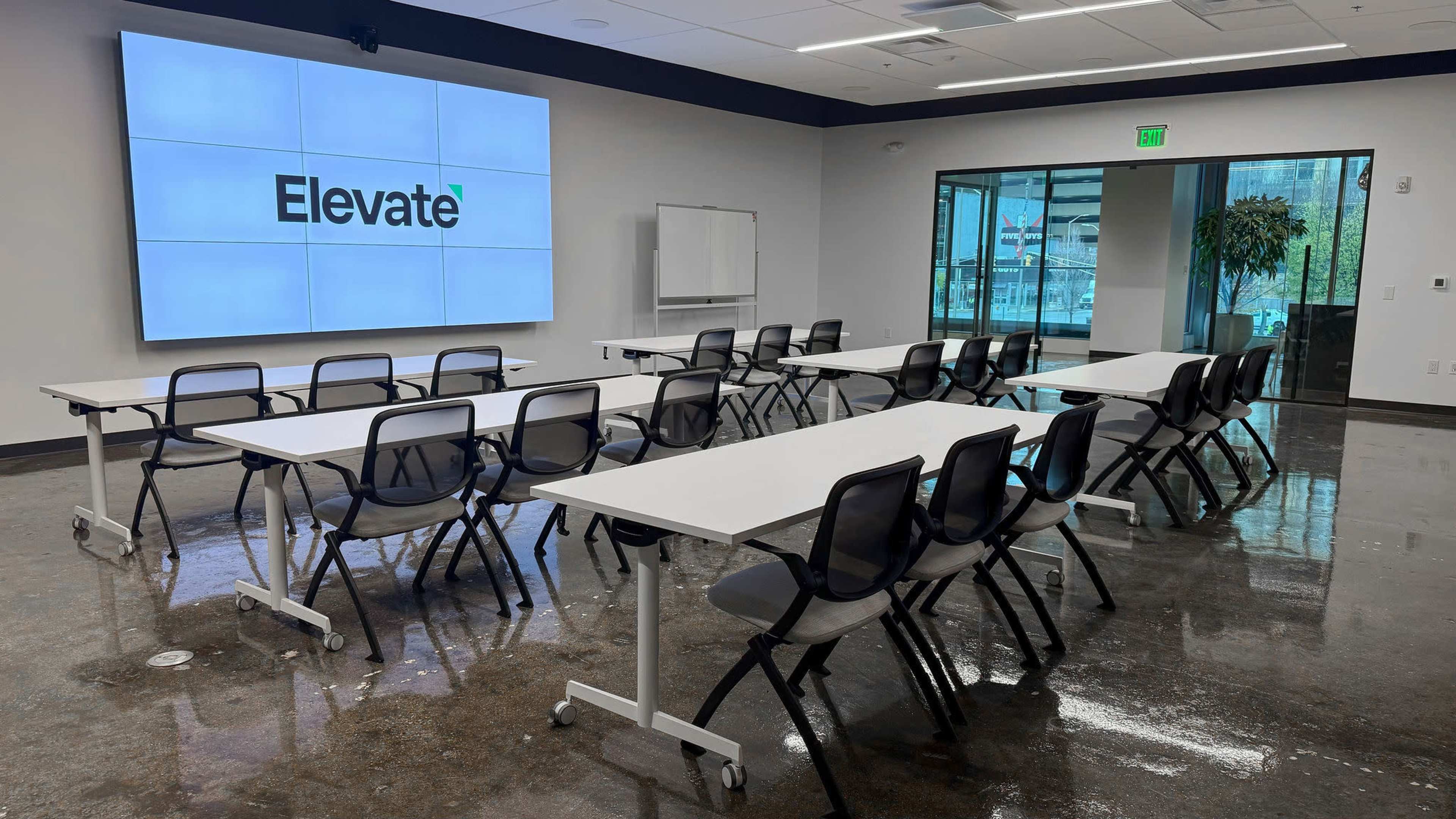 The room features a series of arranged tables and chairs facing a large screen displaying the word "Elevate."