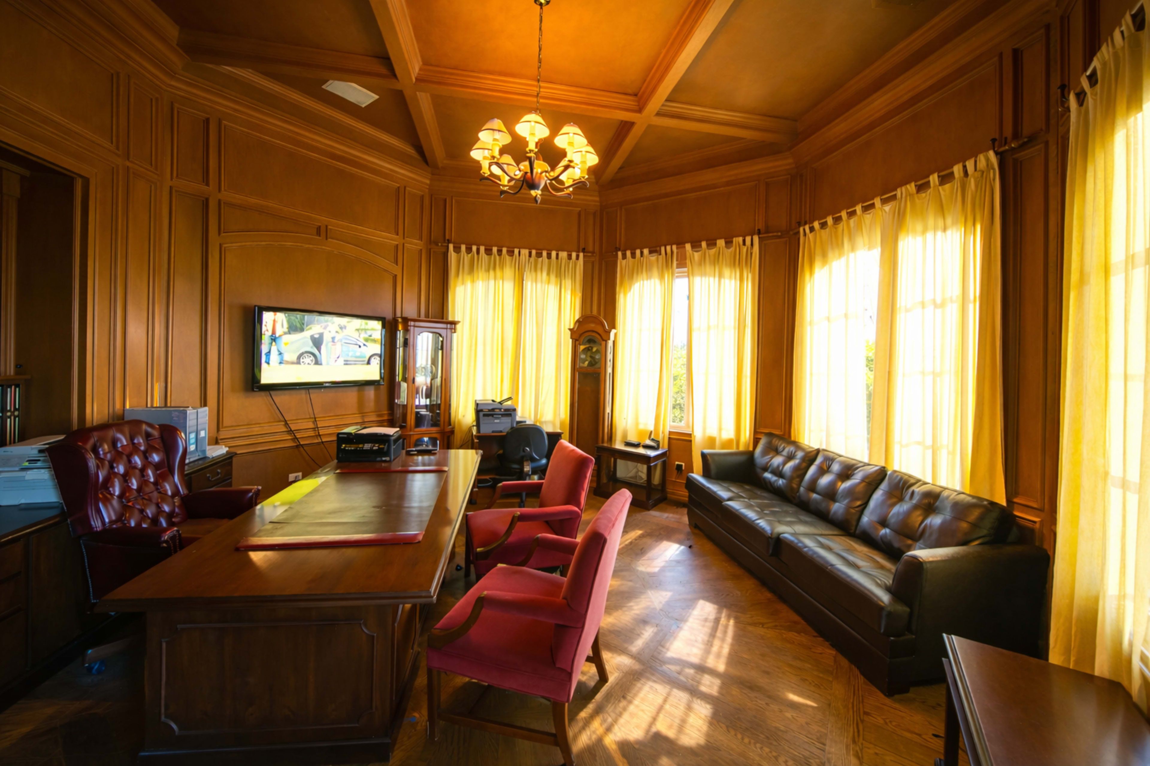 The image shows a wooden-paneled office with a desk, two red chairs, a leather sofa, and large windows dressed with yellow curtains.