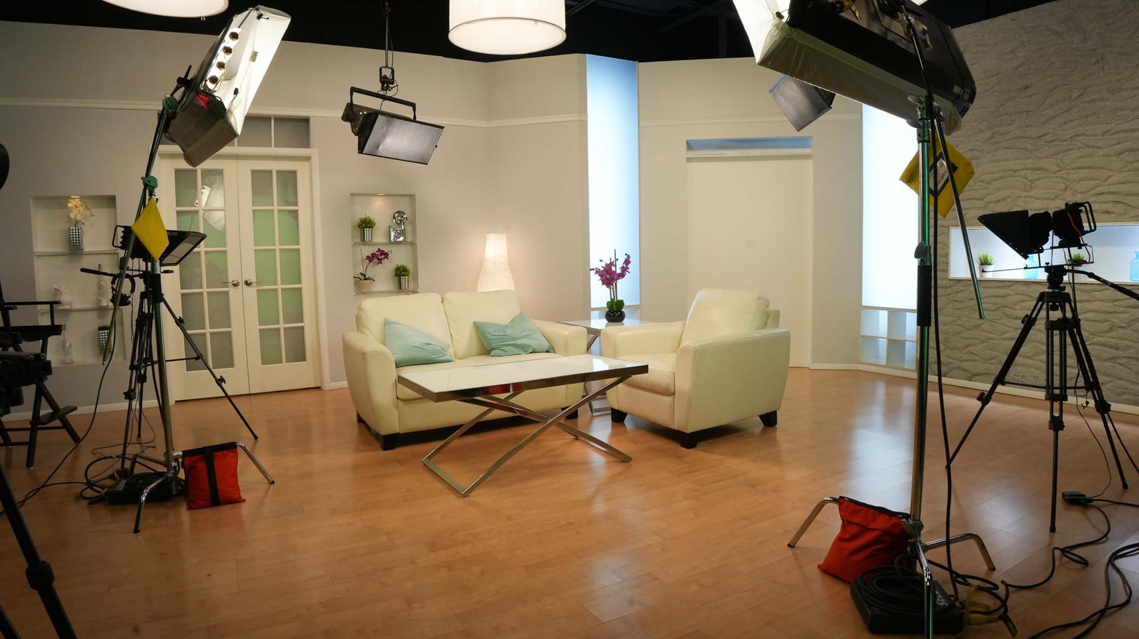 A well-lit television studio features a white sofa and armchair, a glass coffee table, and multiple camera setups.