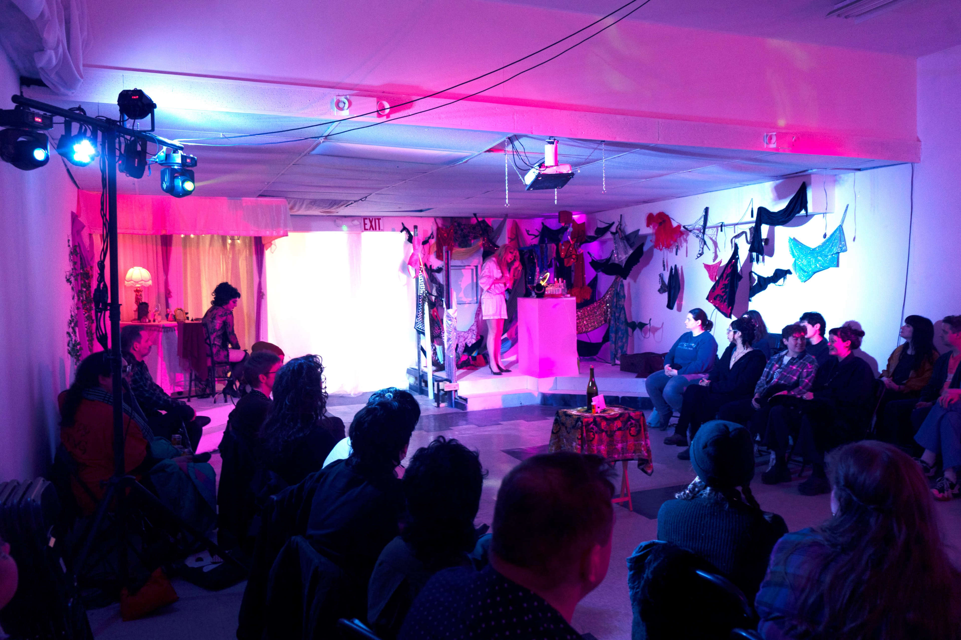 A group of people sits in a dimly lit space with colorful lighting, watching a performance in front of a stage adorned with various costumes and decorations.