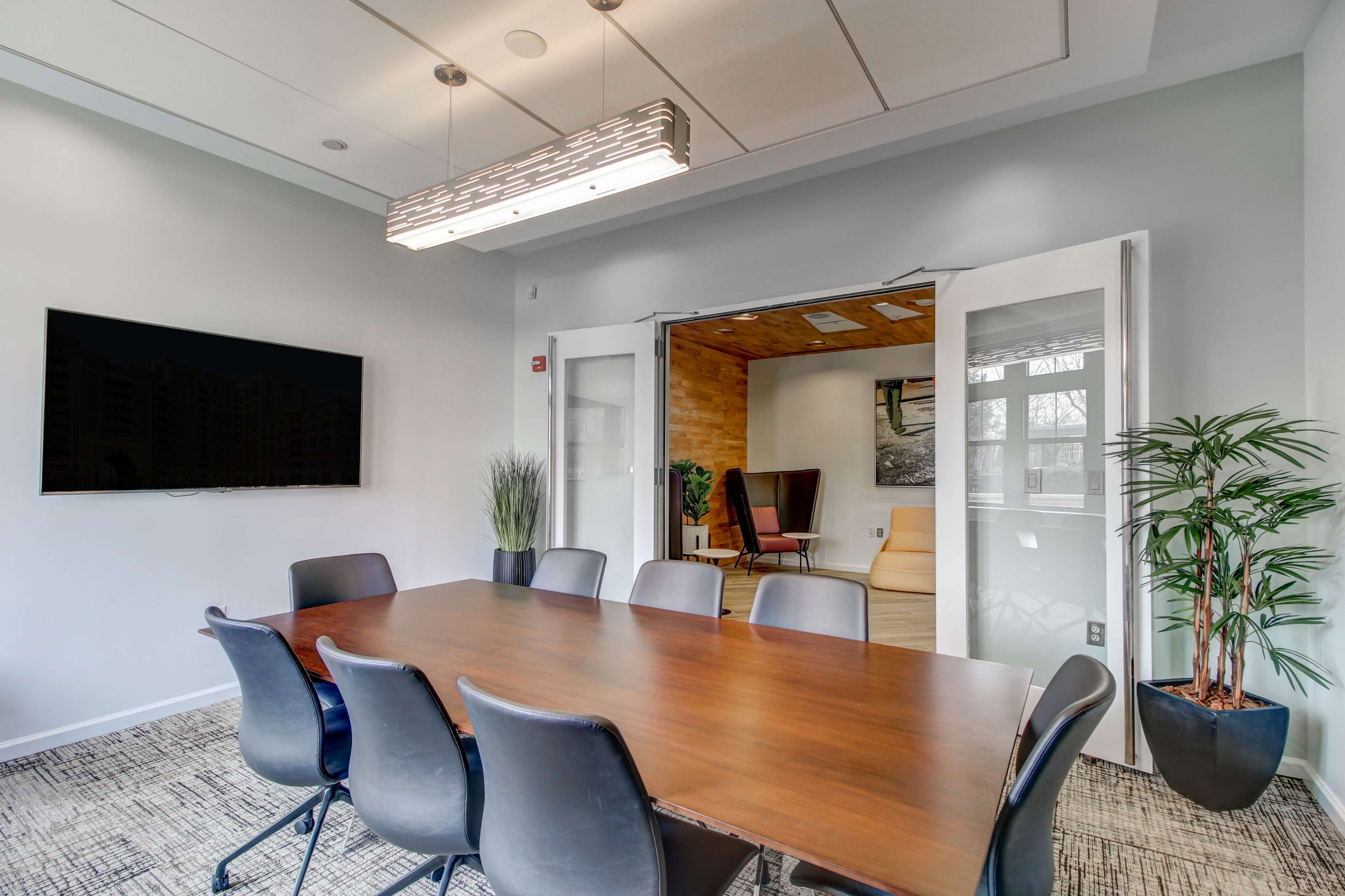 A modern conference room features a long wooden table surrounded by black chairs, with a wall-mounted TV and glass doors leading to an adjoining room.