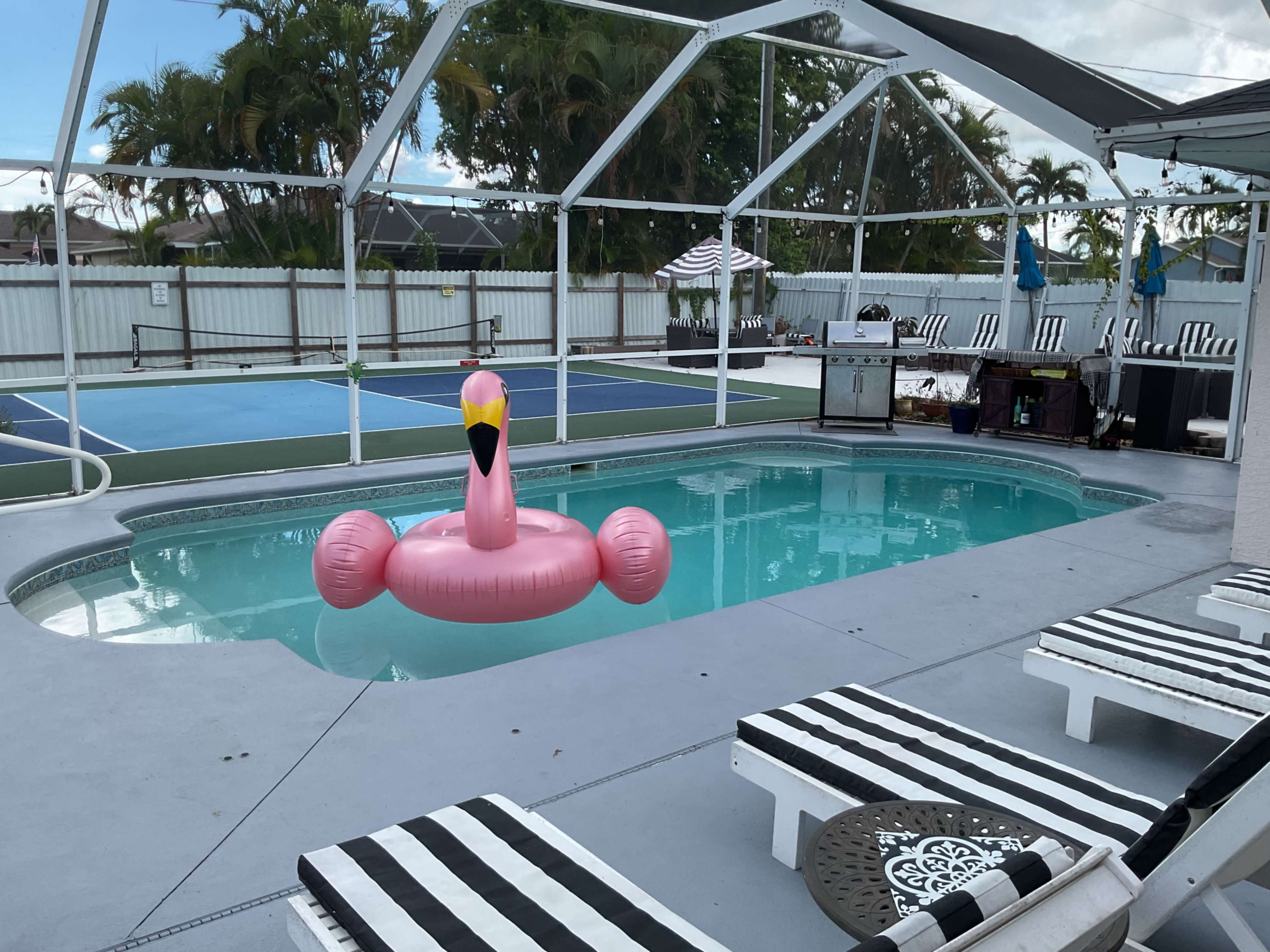 A pink flamingo float sits in a swimming pool surrounded by lounge chairs and a tennis court in the background.