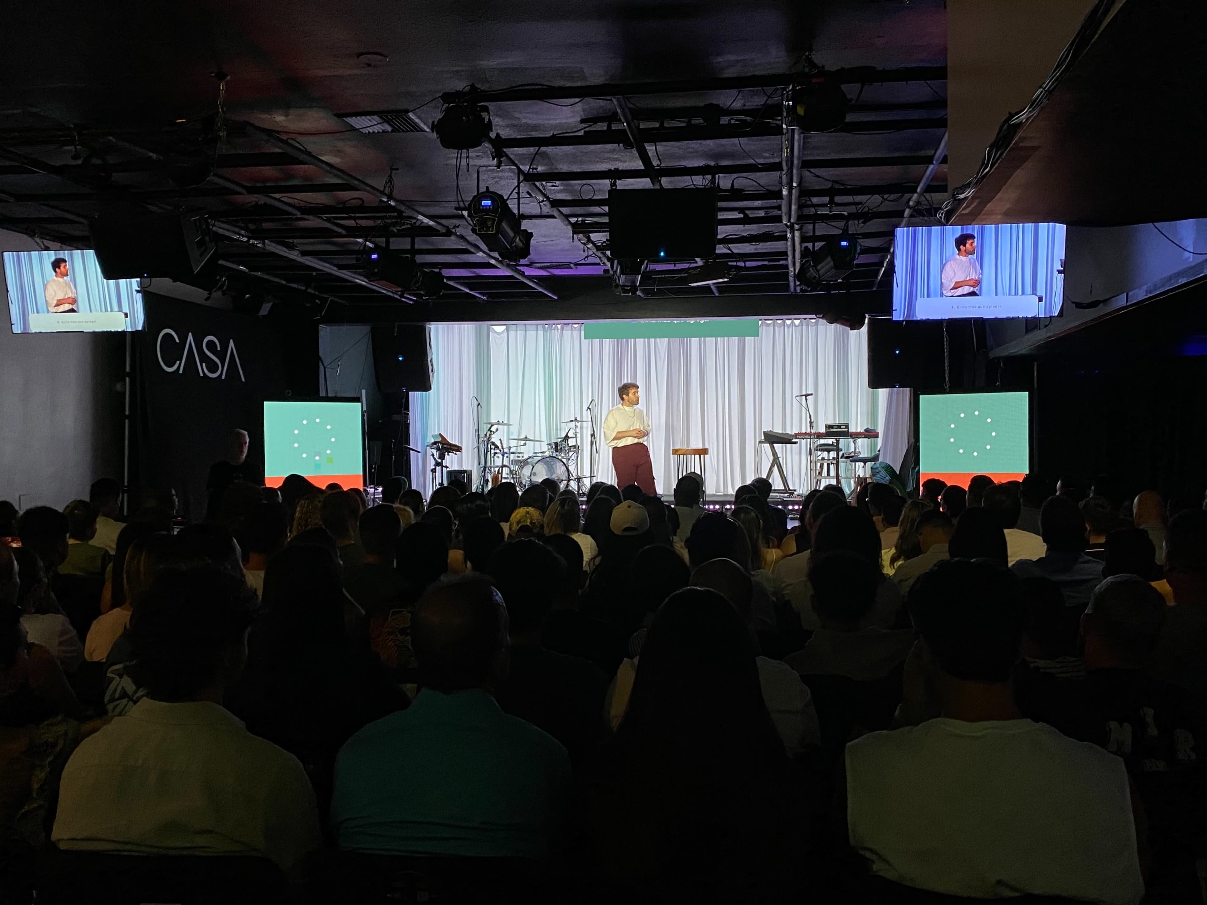 A speaker stands on a stage in front of an audience, with screens displaying visuals and musical instruments arranged in the background.