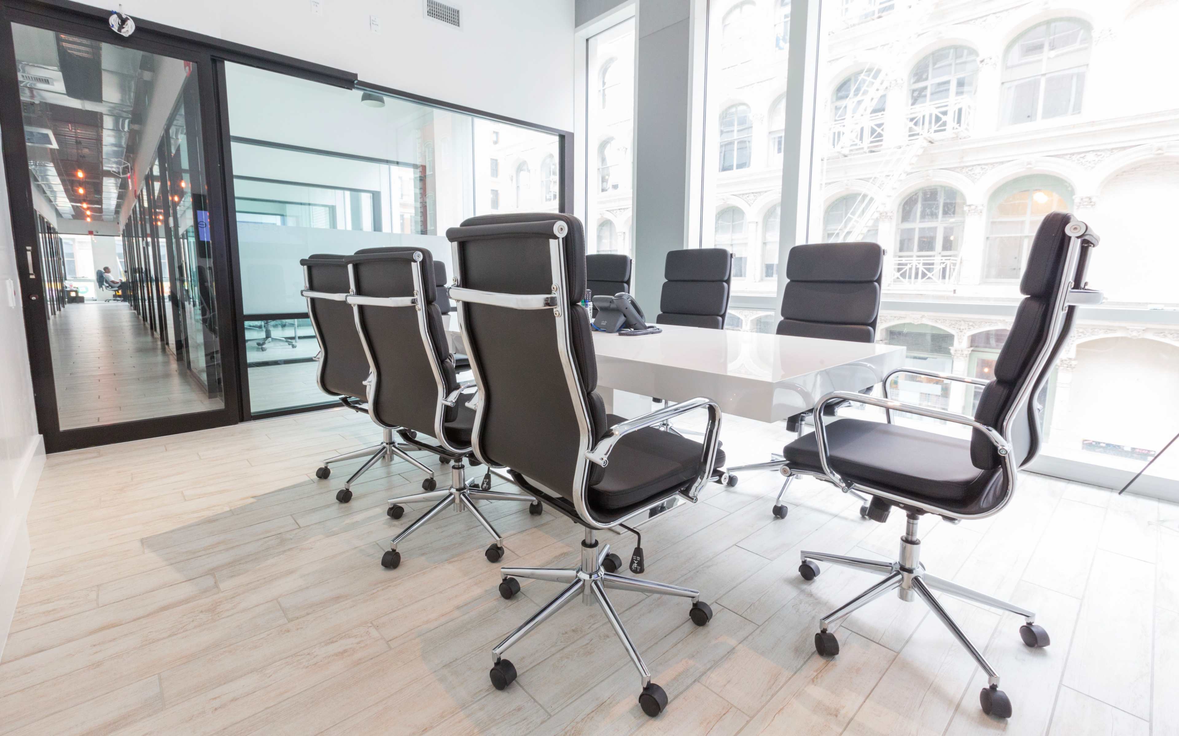A modern conference room features a long white table surrounded by black ergonomic chairs, with large windows offering a view of an exterior building facade.