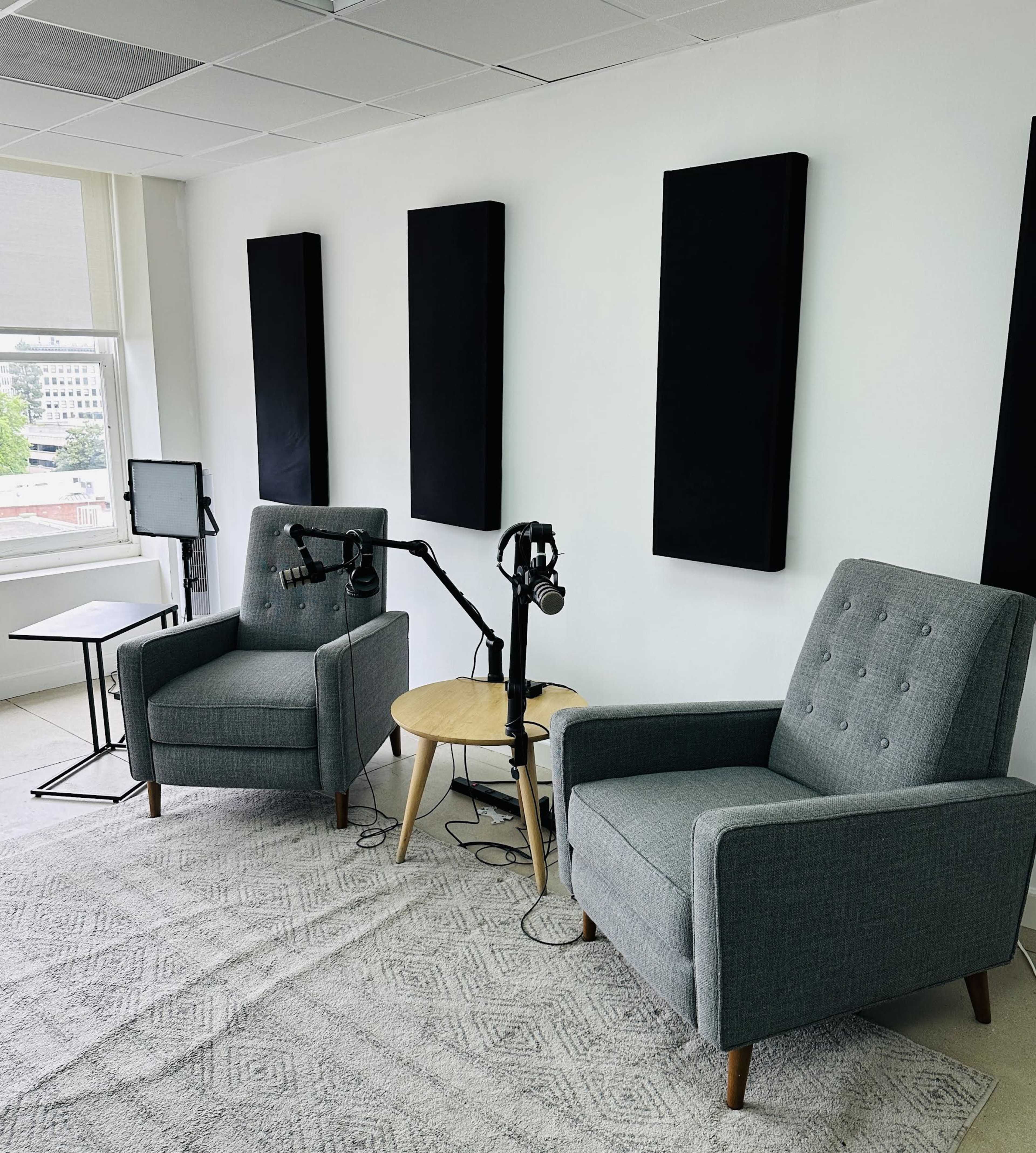 A recording studio with two gray armchairs, a small round table, and microphone stands positioned between them, set against a backdrop of sound-absorbing panels on the walls.