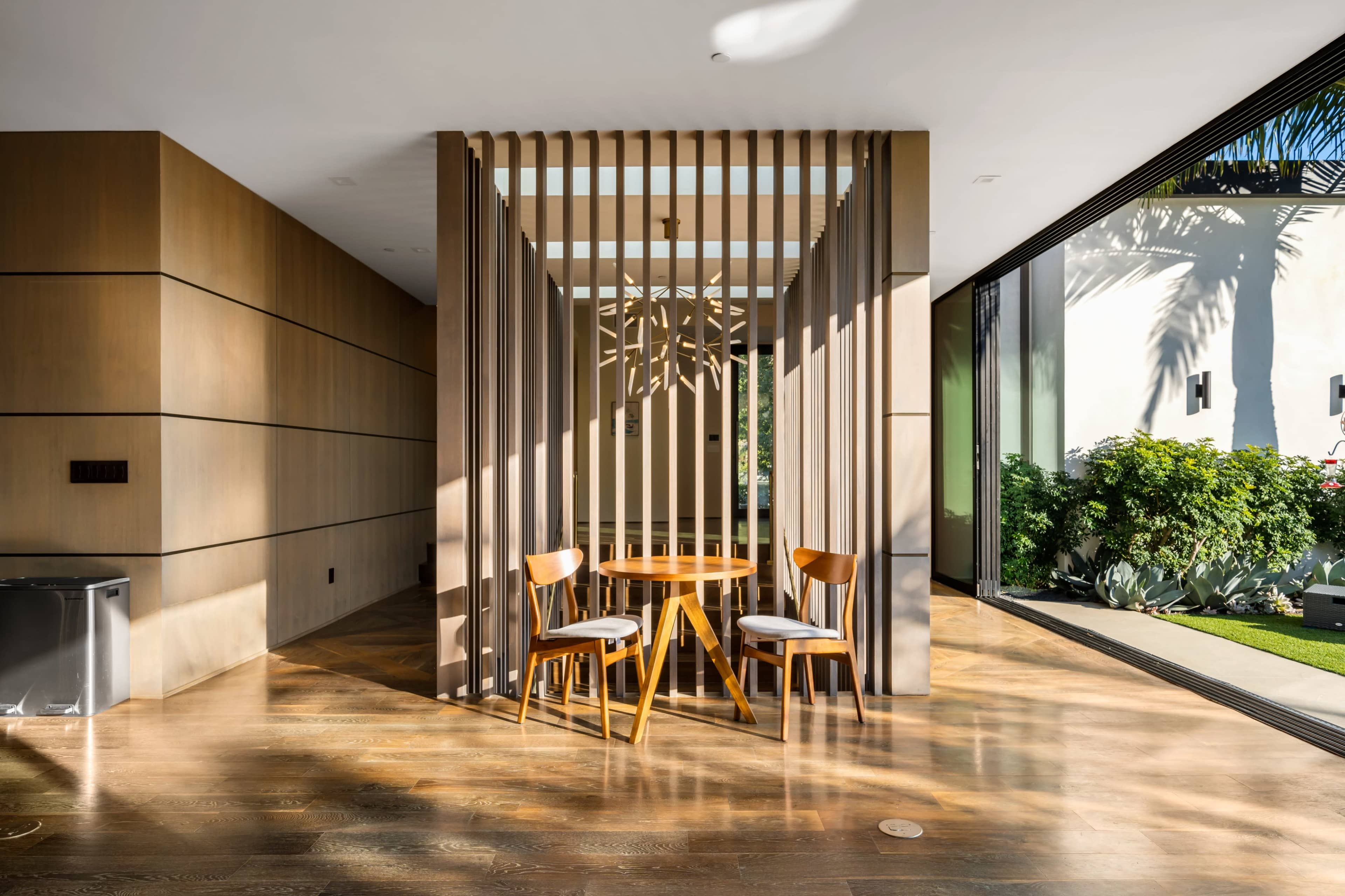 A modern interior space featuring a circular dining table surrounded by three wooden chairs, divided by a slatted wooden partition.