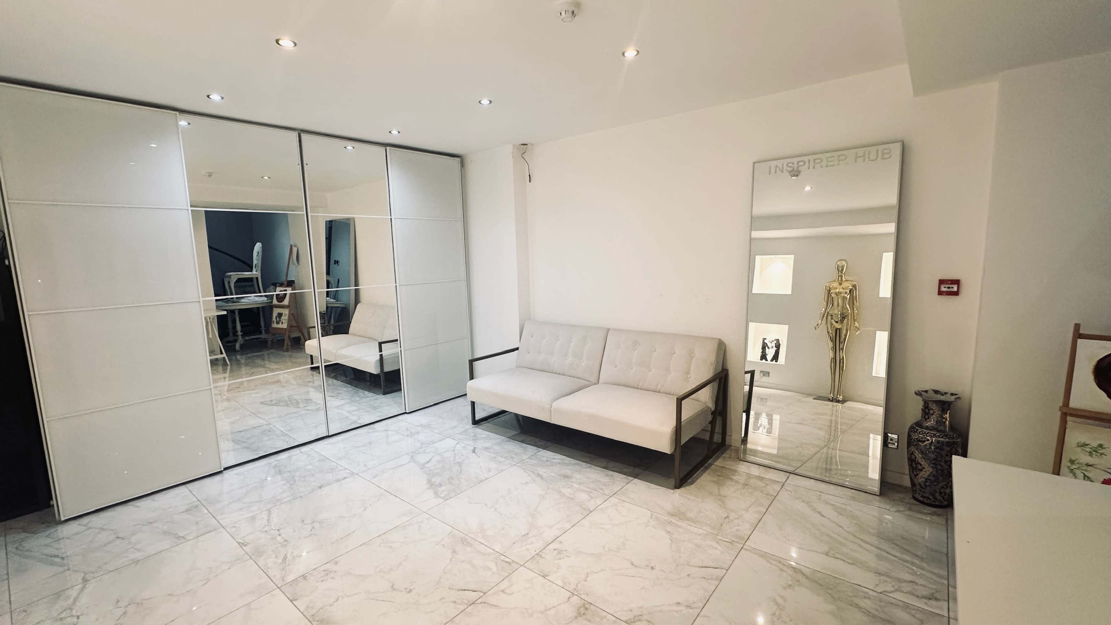 The image depicts a modern, minimalist interior with a white sofa, mirrored wardrobe doors, and a decorative mirror reflecting a skeleton figurine.