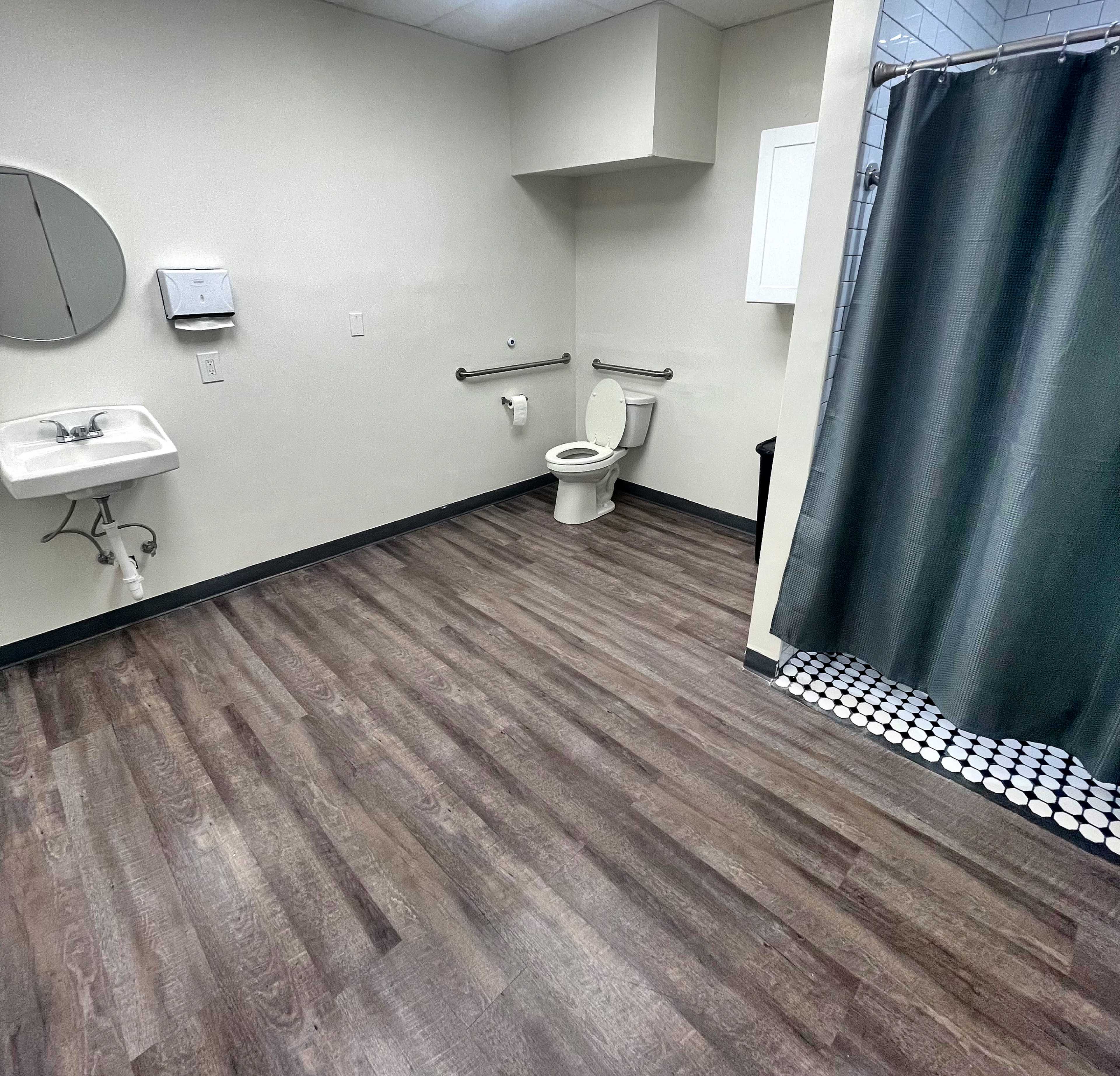 The image shows a clean, unoccupied restroom with a toilet, sink, and a shower area separated by a curtain.