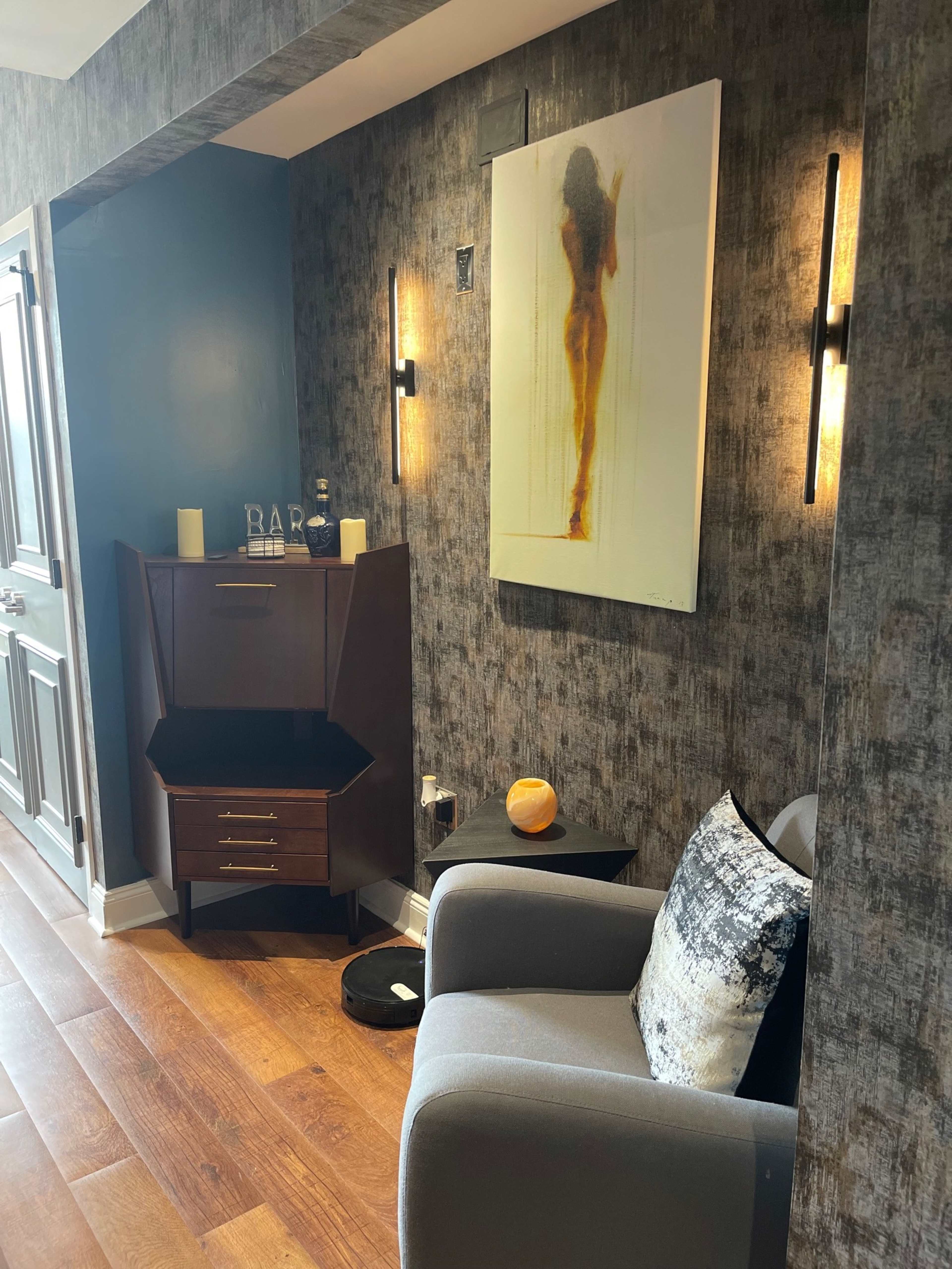 A modern interior corner featuring a gray armchair, a dark wood cabinet, wall sconces, and an abstract painting.