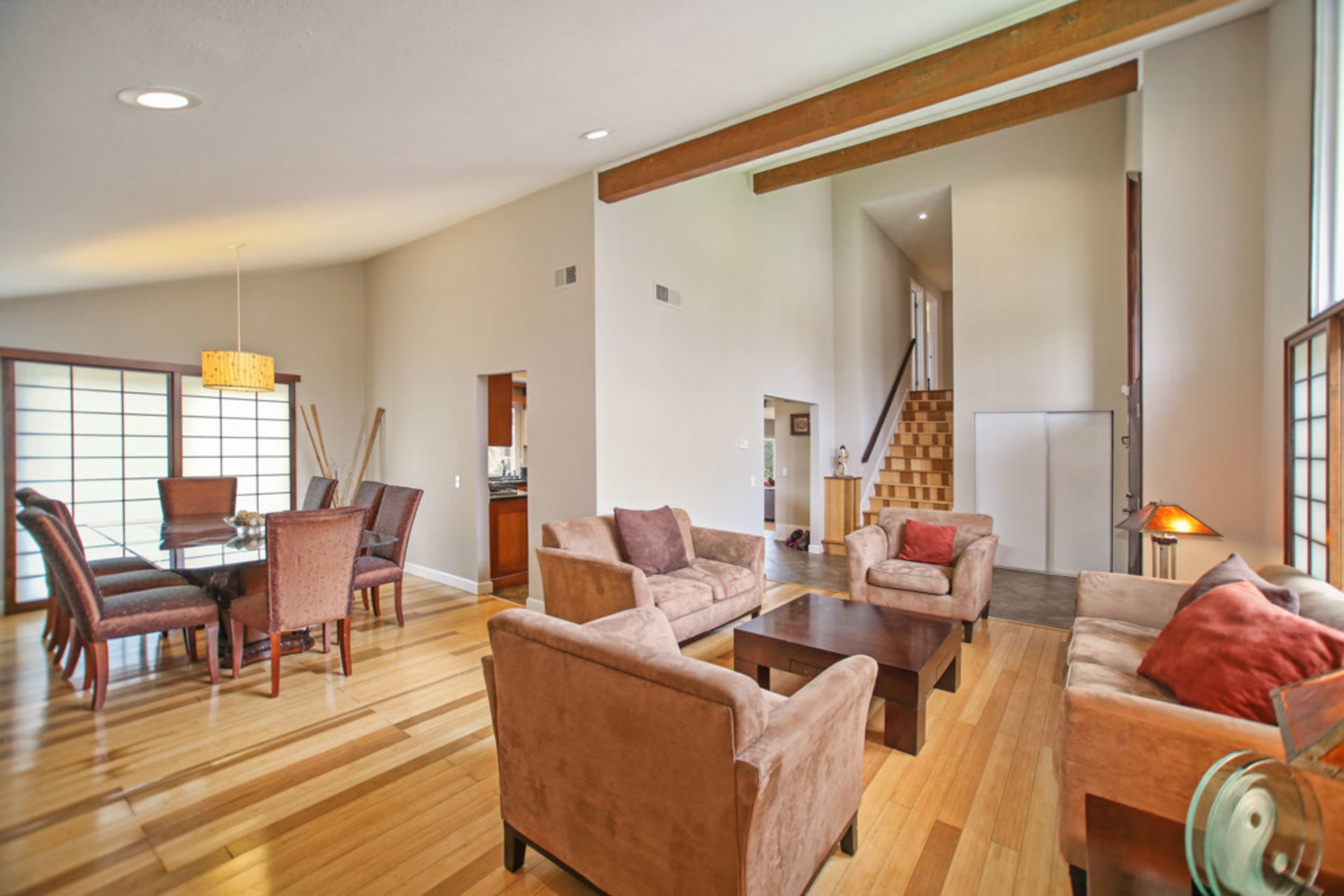 The image shows a spacious living area with a dining table, several seating arrangements, and a staircase leading to an upper level.