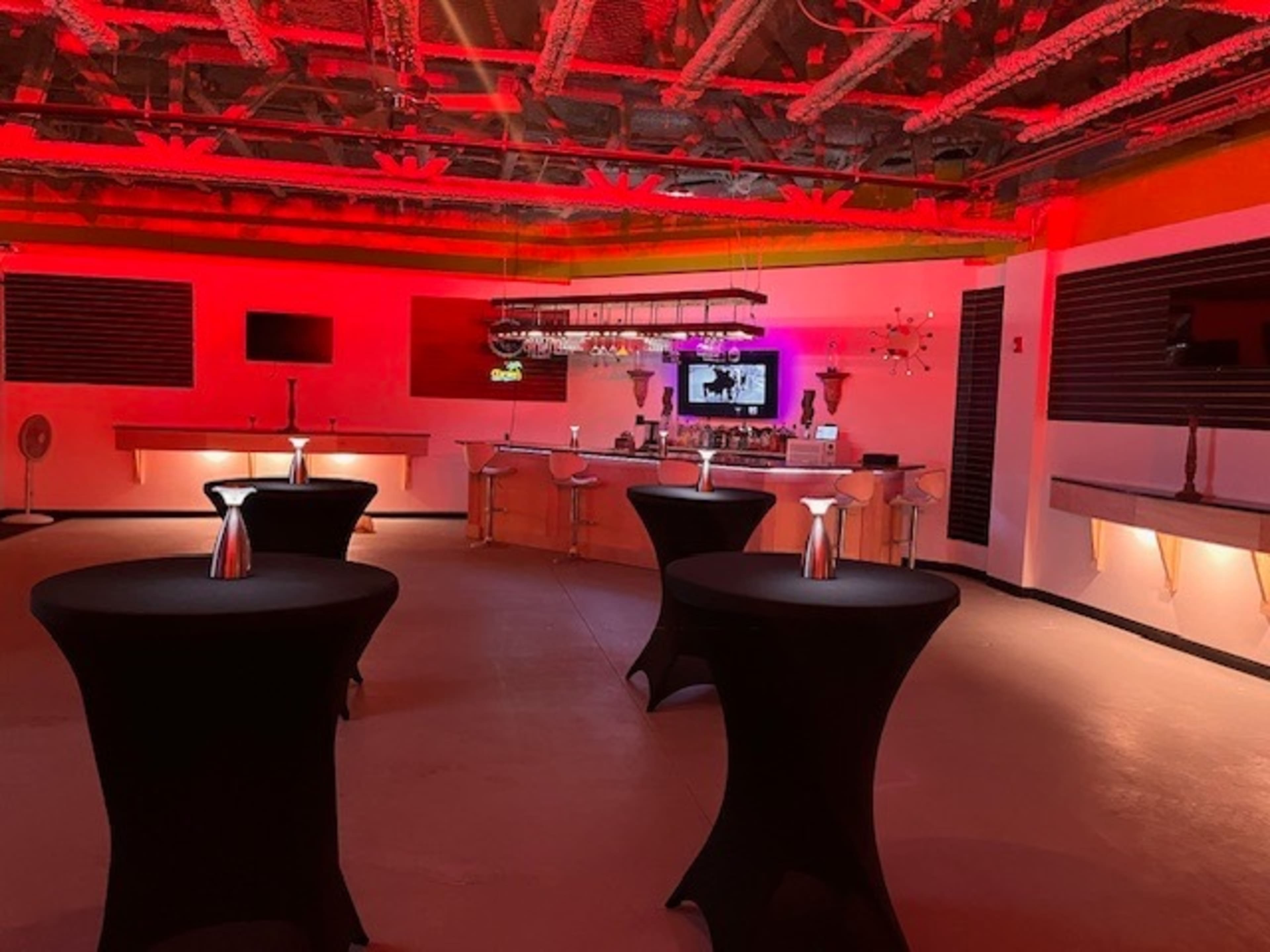 A dimly lit event space features high tables with black cloths, a bar area, and red lighting on the walls and ceiling.