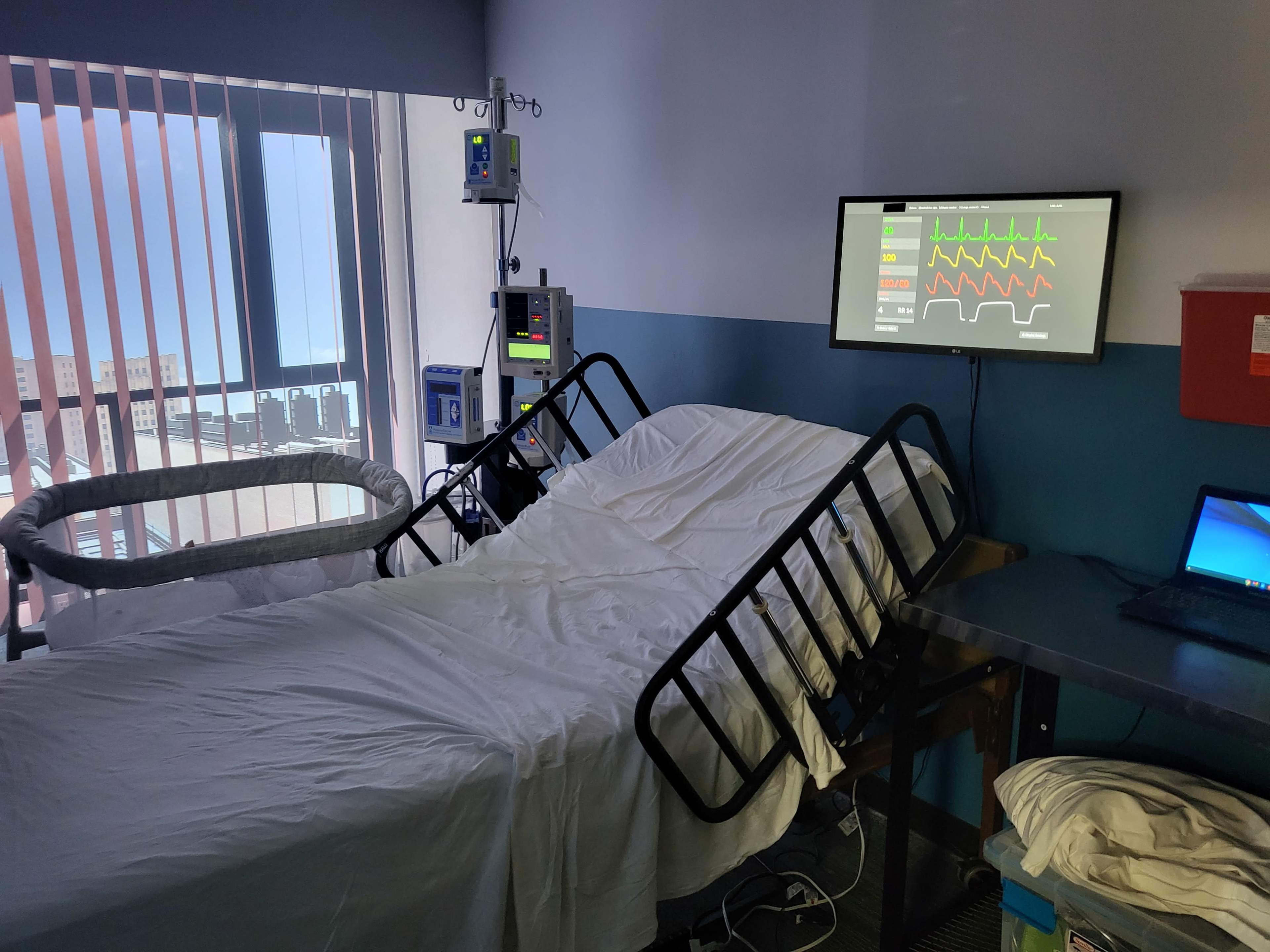 The room contains a hospital bed, medical monitoring equipment, and a laptop, with a large window letting in natural light.