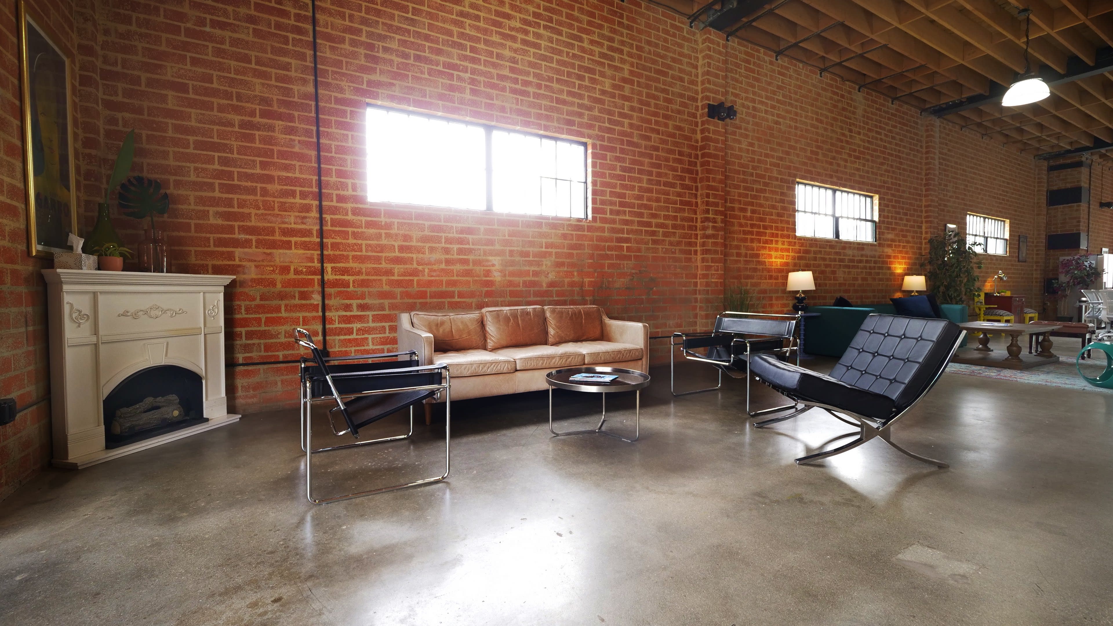The interior features a spacious room with exposed brick walls, a couch, two modern chairs, a coffee table, and large windows letting in natural light.