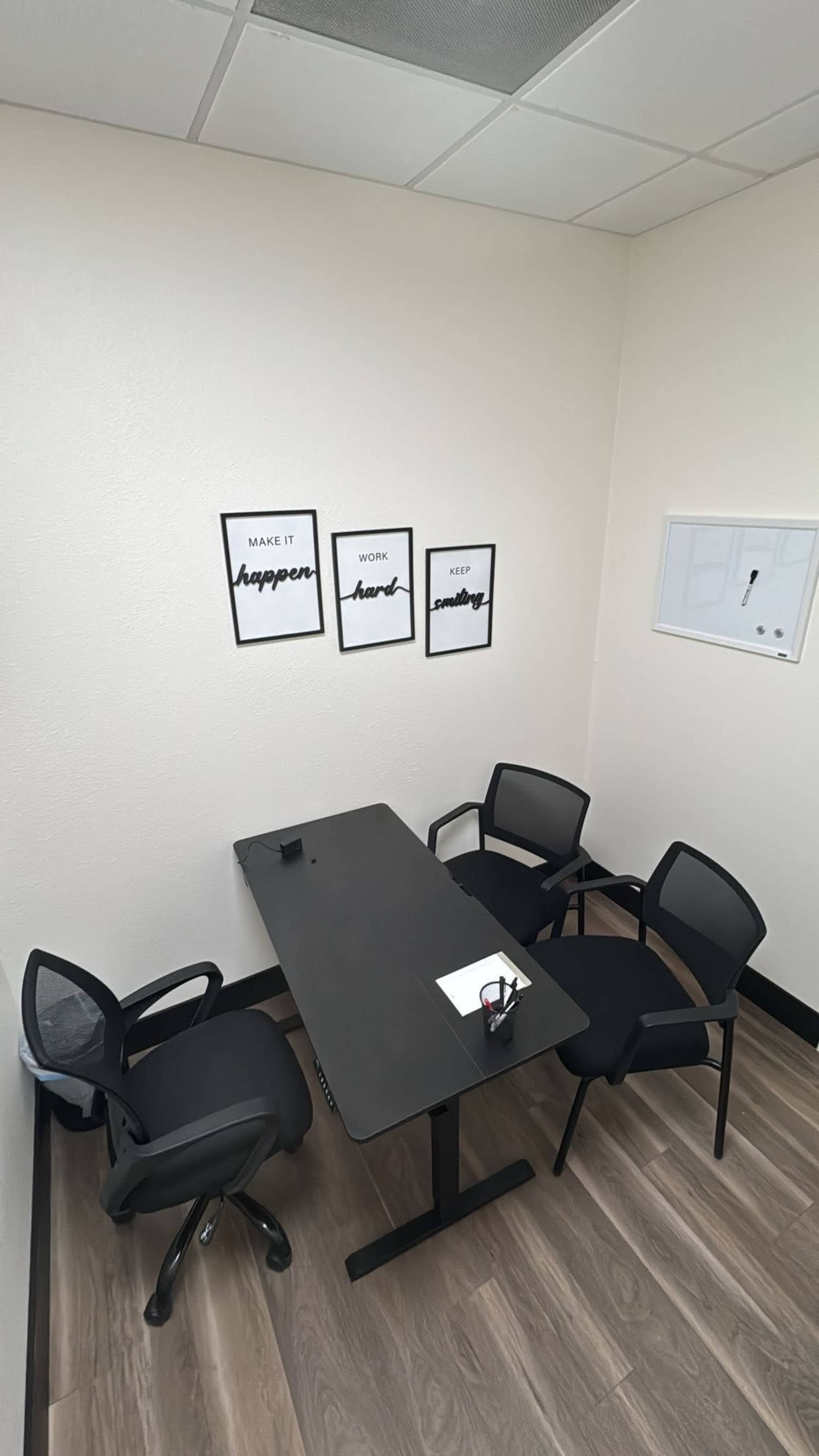 The image shows a small meeting room with a rectangular table and four black chairs, featuring motivational posters on the wall.